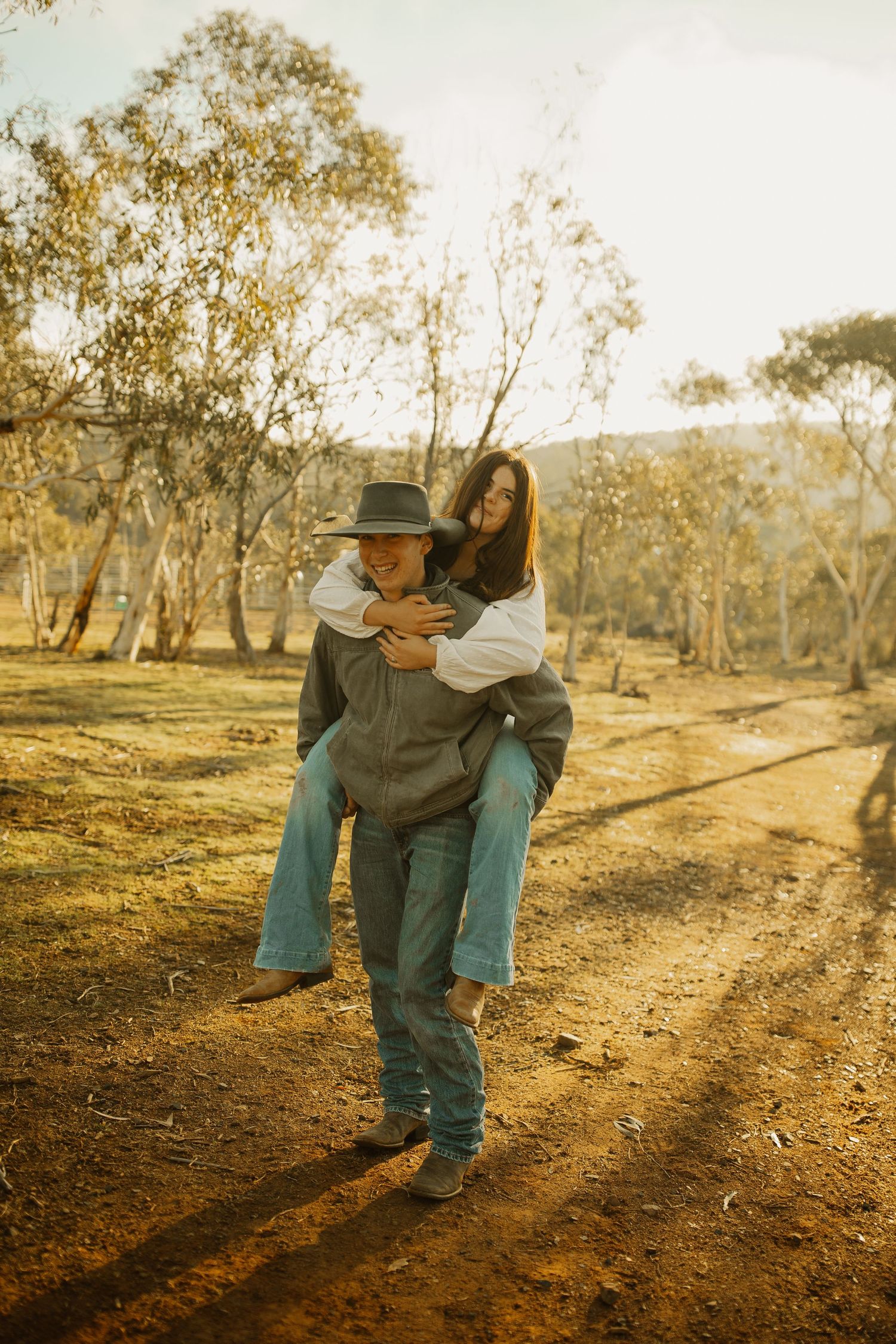 A couple embraces on a sunlit dirt path through an Australian bush landscape in warm golden tones.