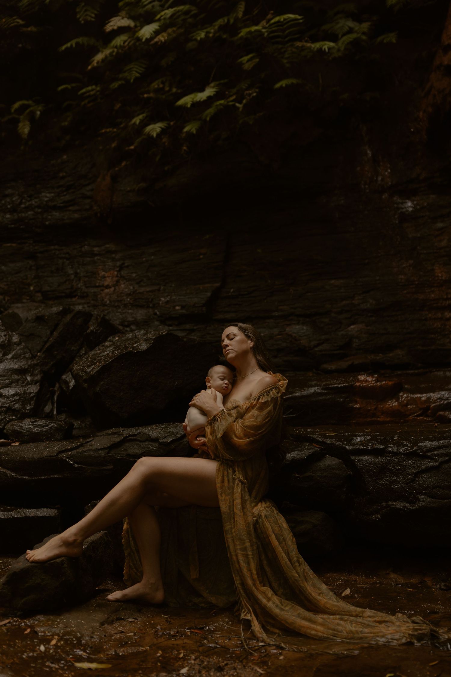 An ethereal scene with a figure in gold dress sitting on rocks beside a dark forest pond holding an infant.