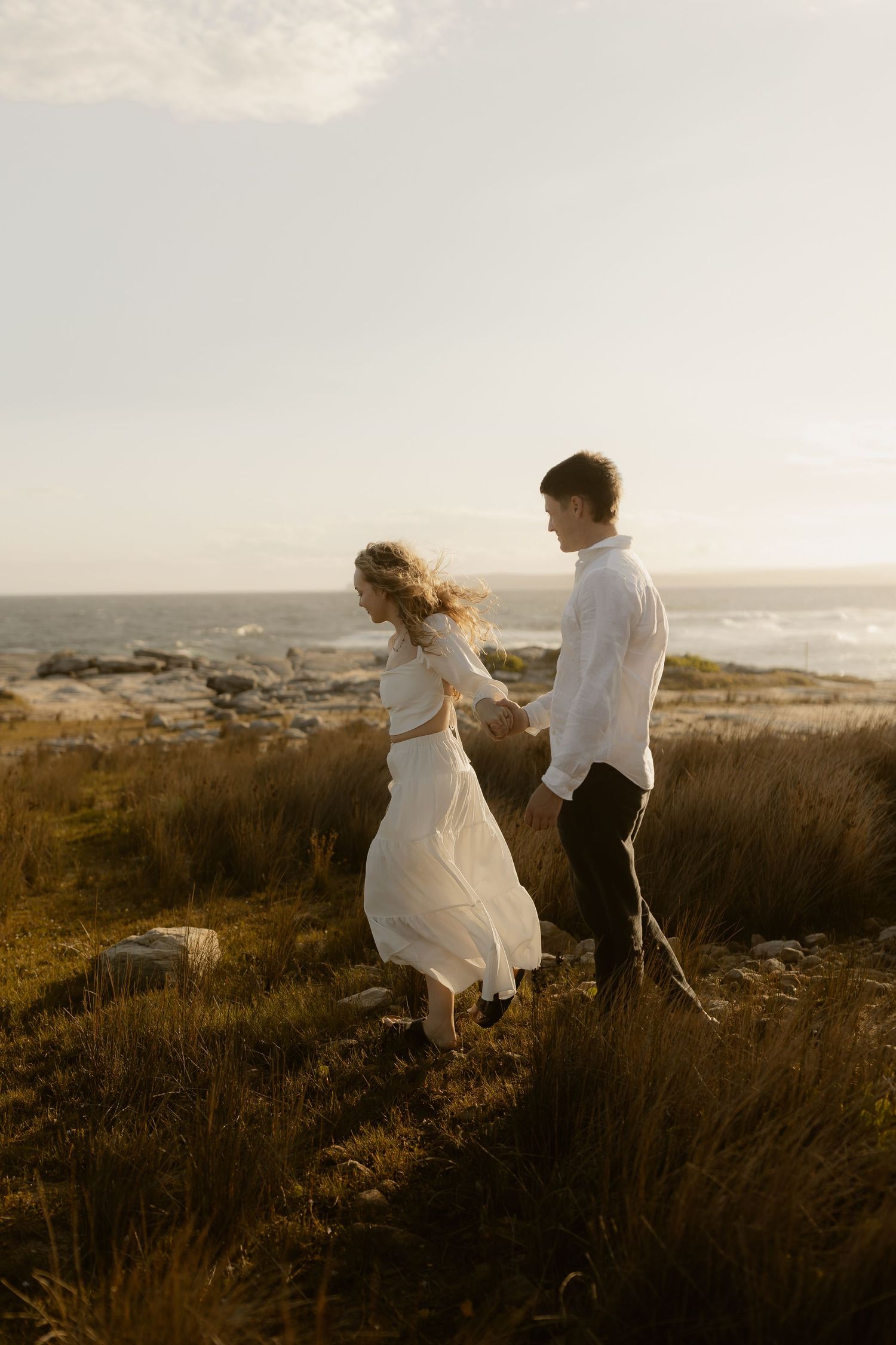Two people in white clothing running through tall grass near the coastline during sunset.