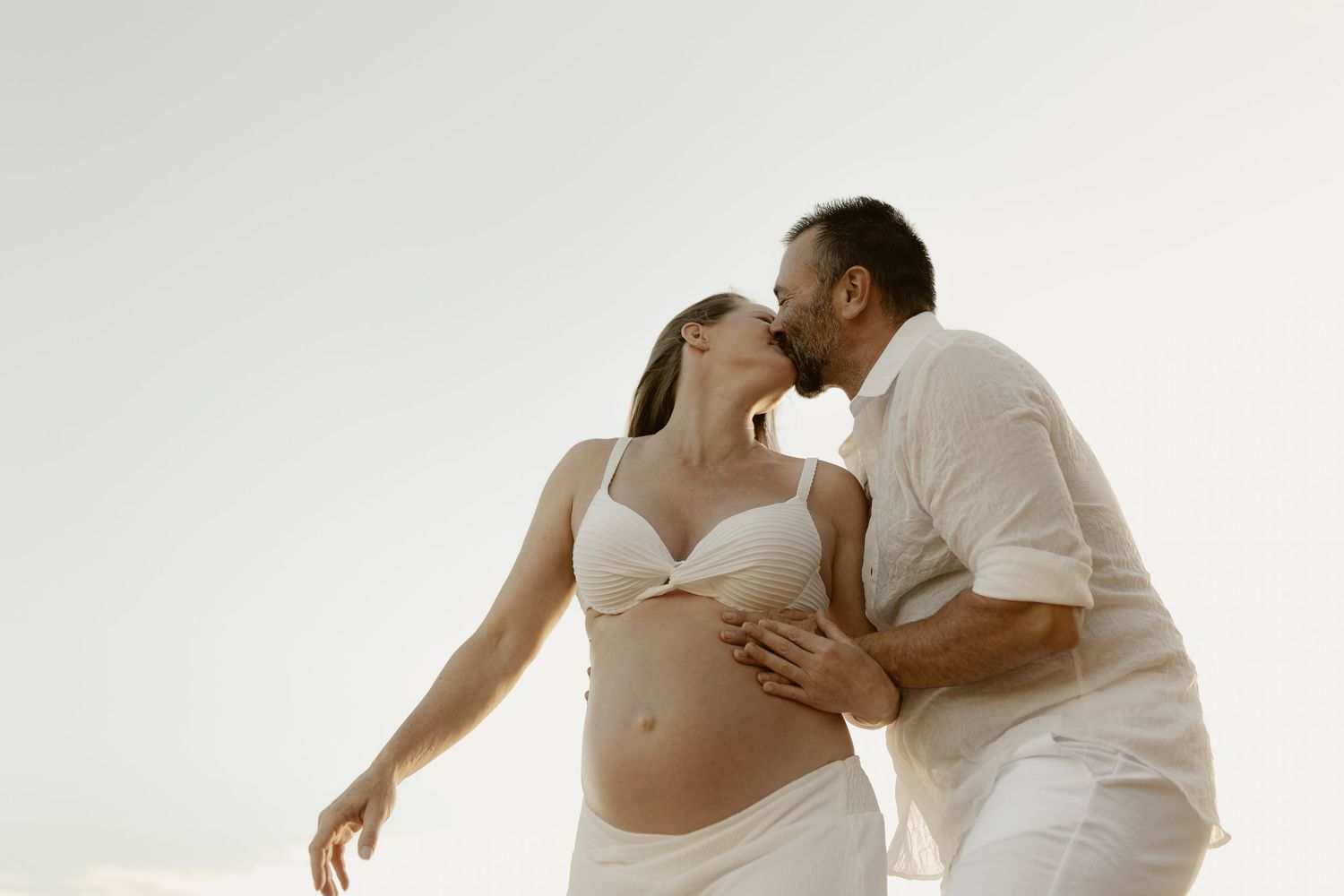 A romantic couple in white clothing embraces and dances together in an elegant pose against a light background.