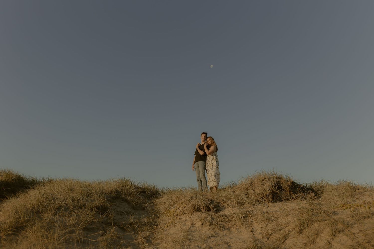 A couple stands together on a sandy hilltop against a dusky blue sky.