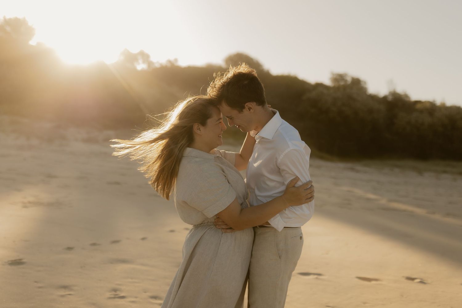 A couple embraces on a sunlit beach during golden hour as their white clothing glows in the warm evening light.