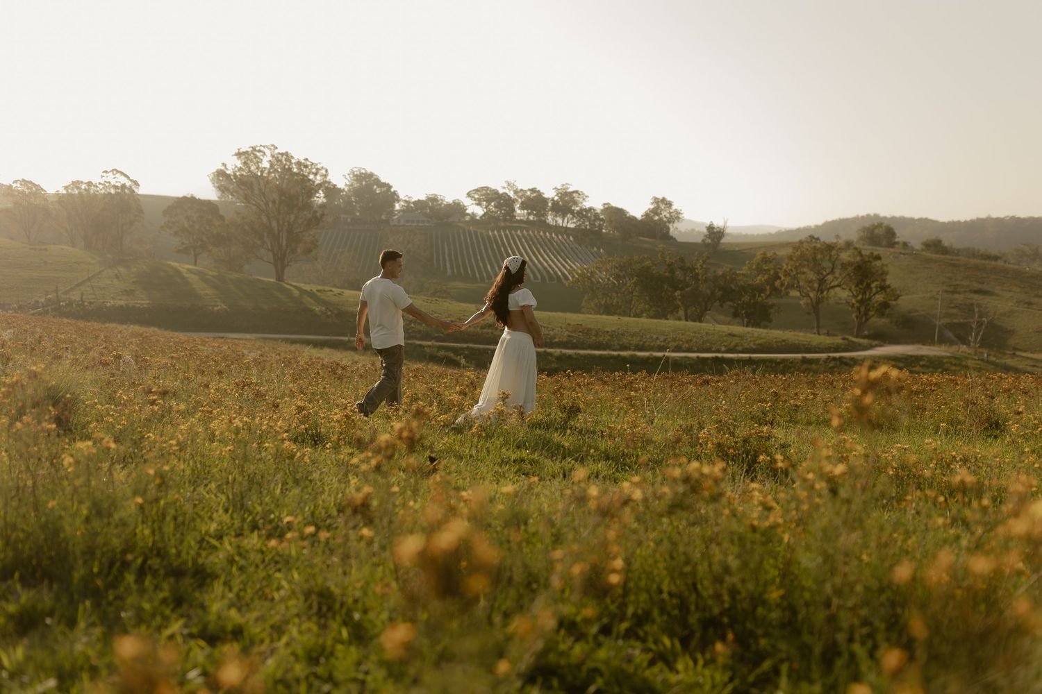 Two people run hand-in-hand through a wildflower field at sunset with rolling hills in the distance.