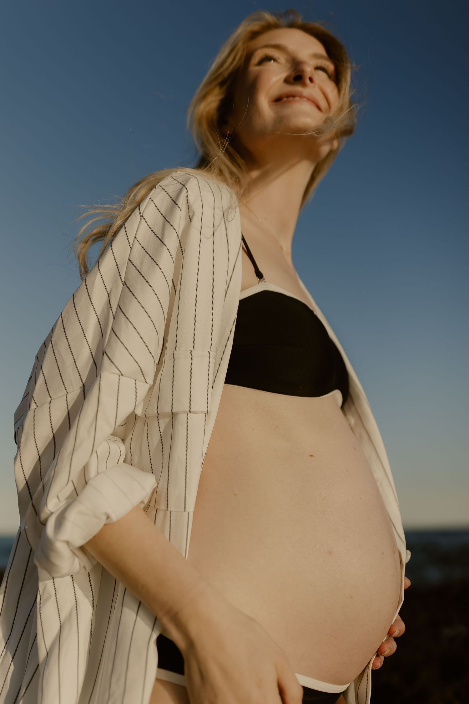 Woman in a white striped blazer poses against a blue sky during golden hour.