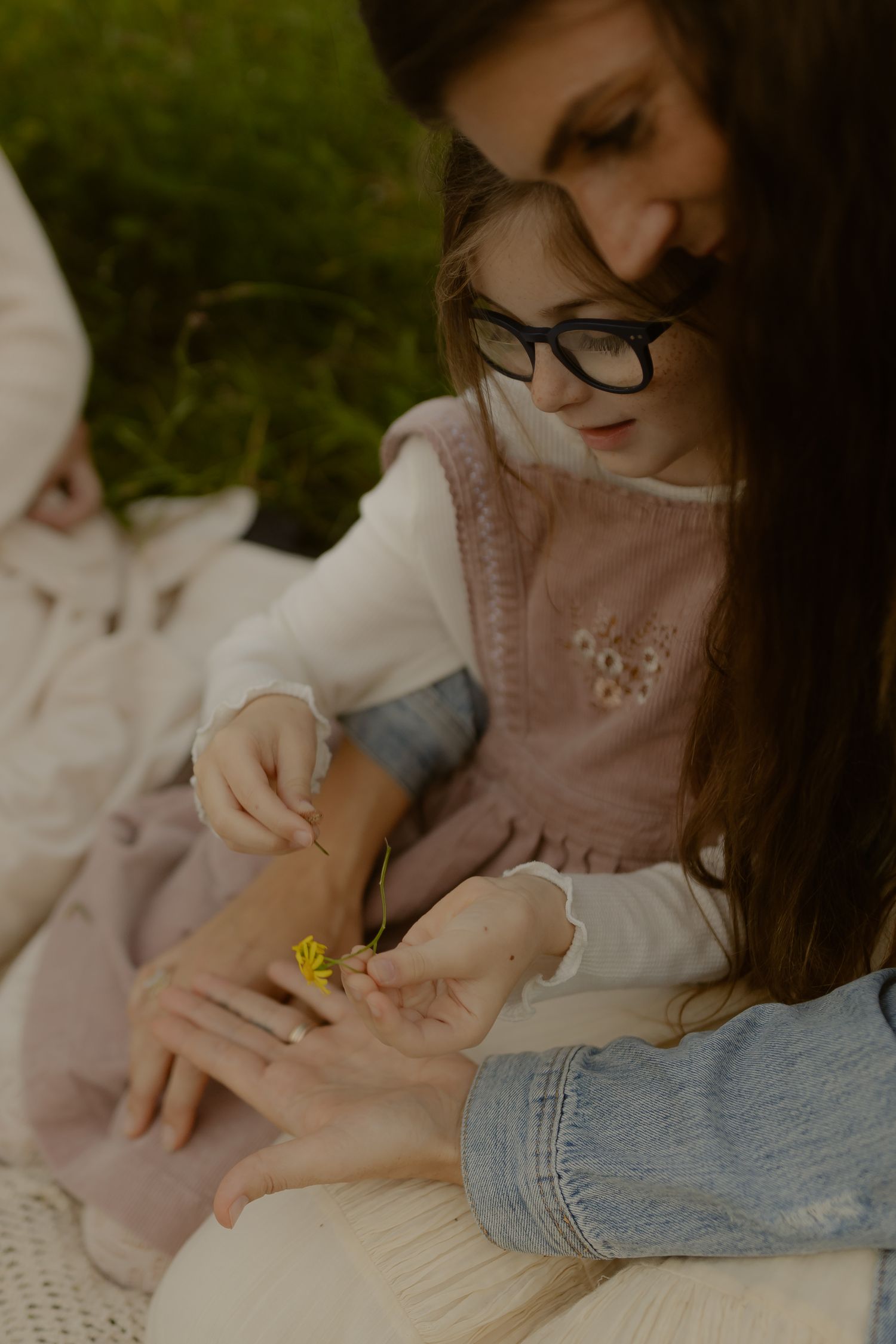 Young girl with glasses examining a small yellow flower with a woman during an outdoor picnic on green grass.