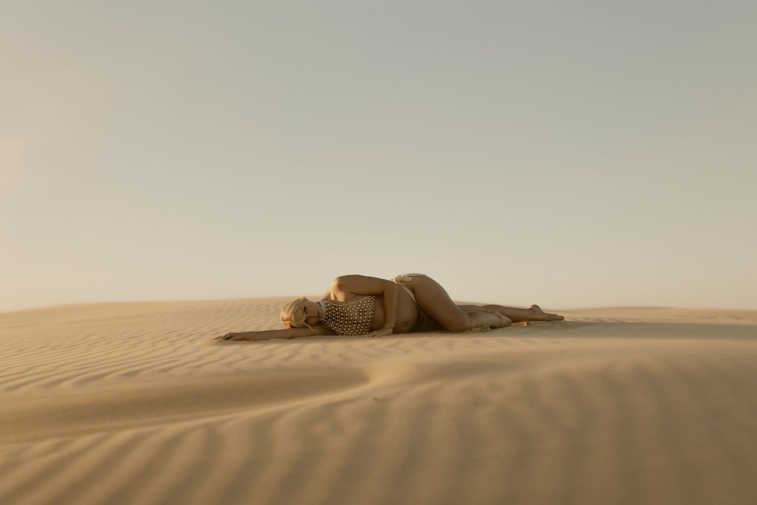 A person lying curled up on golden sand dunes in a vast desert landscape under a hazy sky.