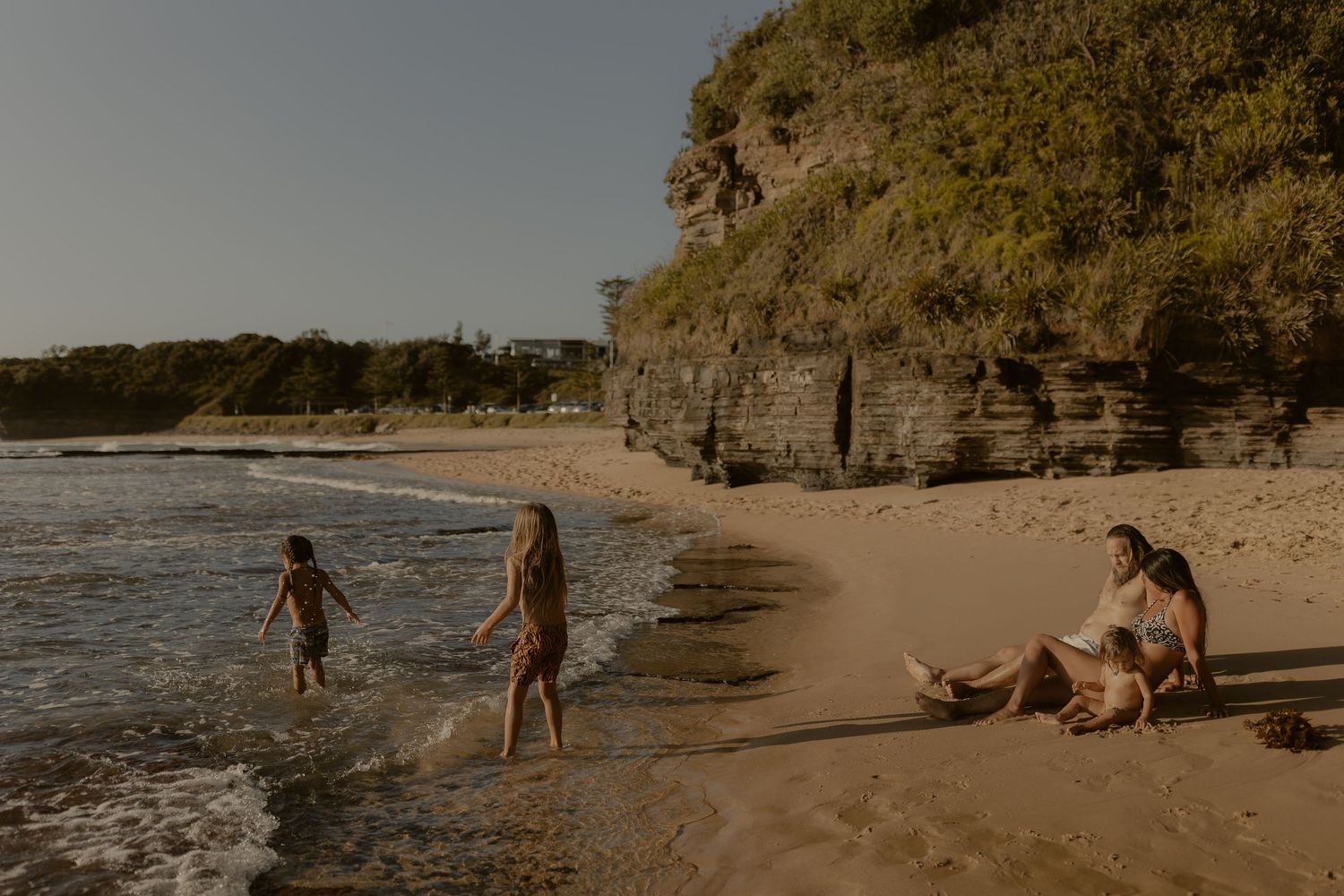 A scenic beach landscape with rocky cliffs and people walking along the shoreline at sunset.