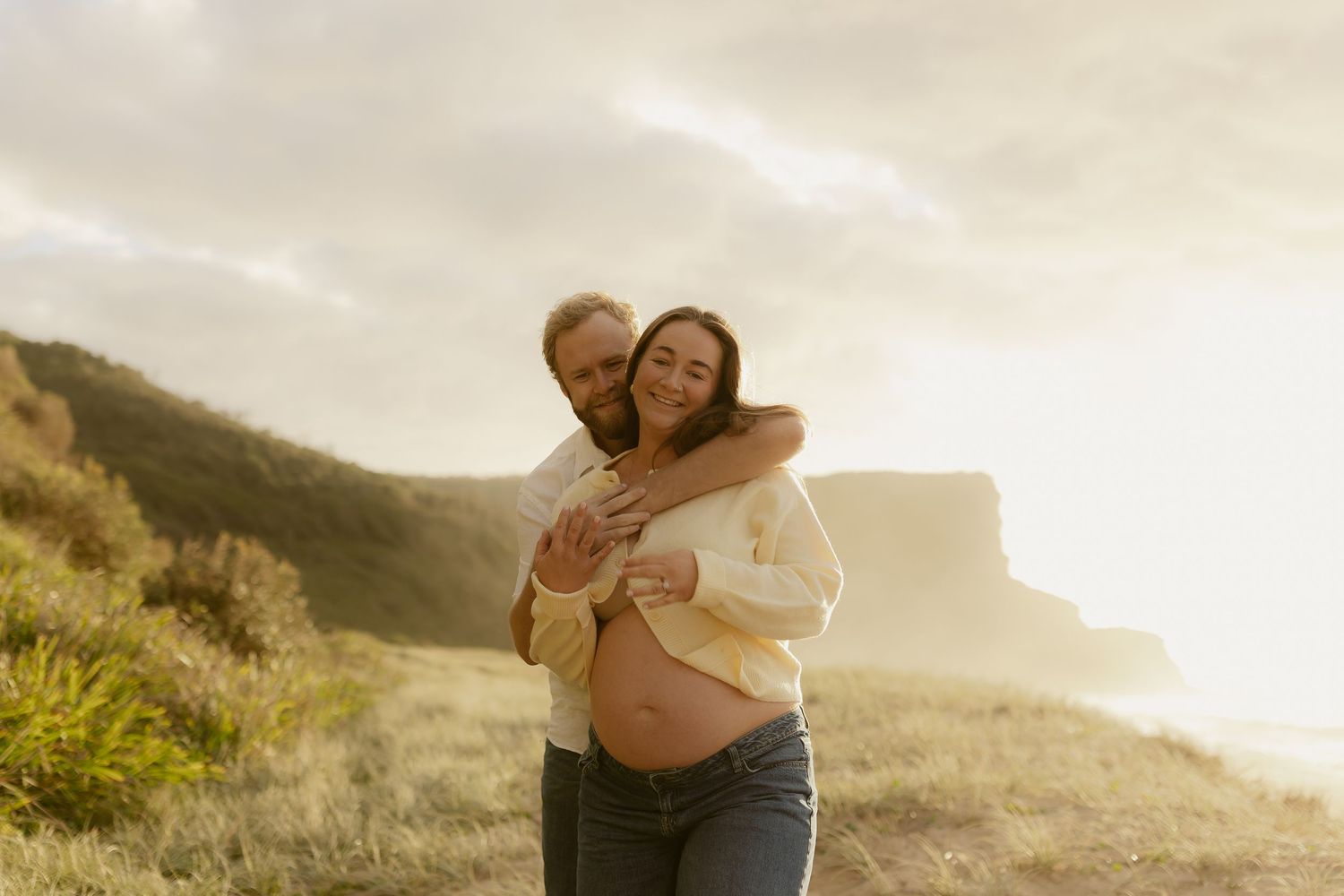 Couple embraces on a scenic hilltop overlooking the ocean at sunset.
