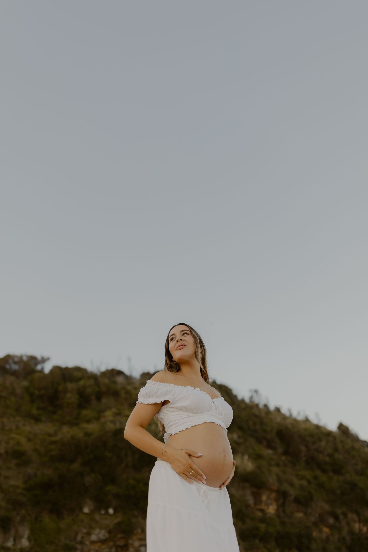 A pregnant woman in a white dress poses against a hillside background during golden hour.