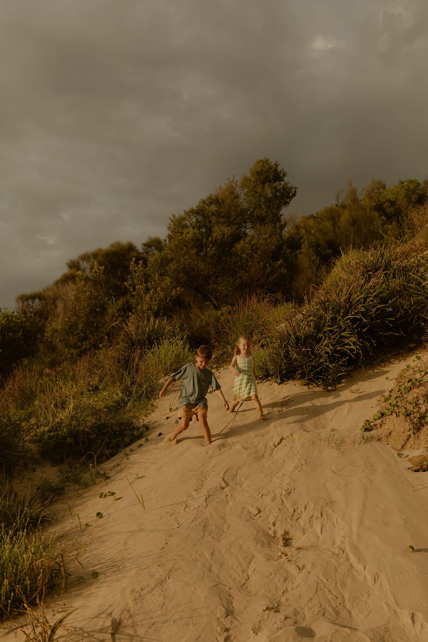 People climbing sandy dunes under dramatic cloudy skies at sunset.