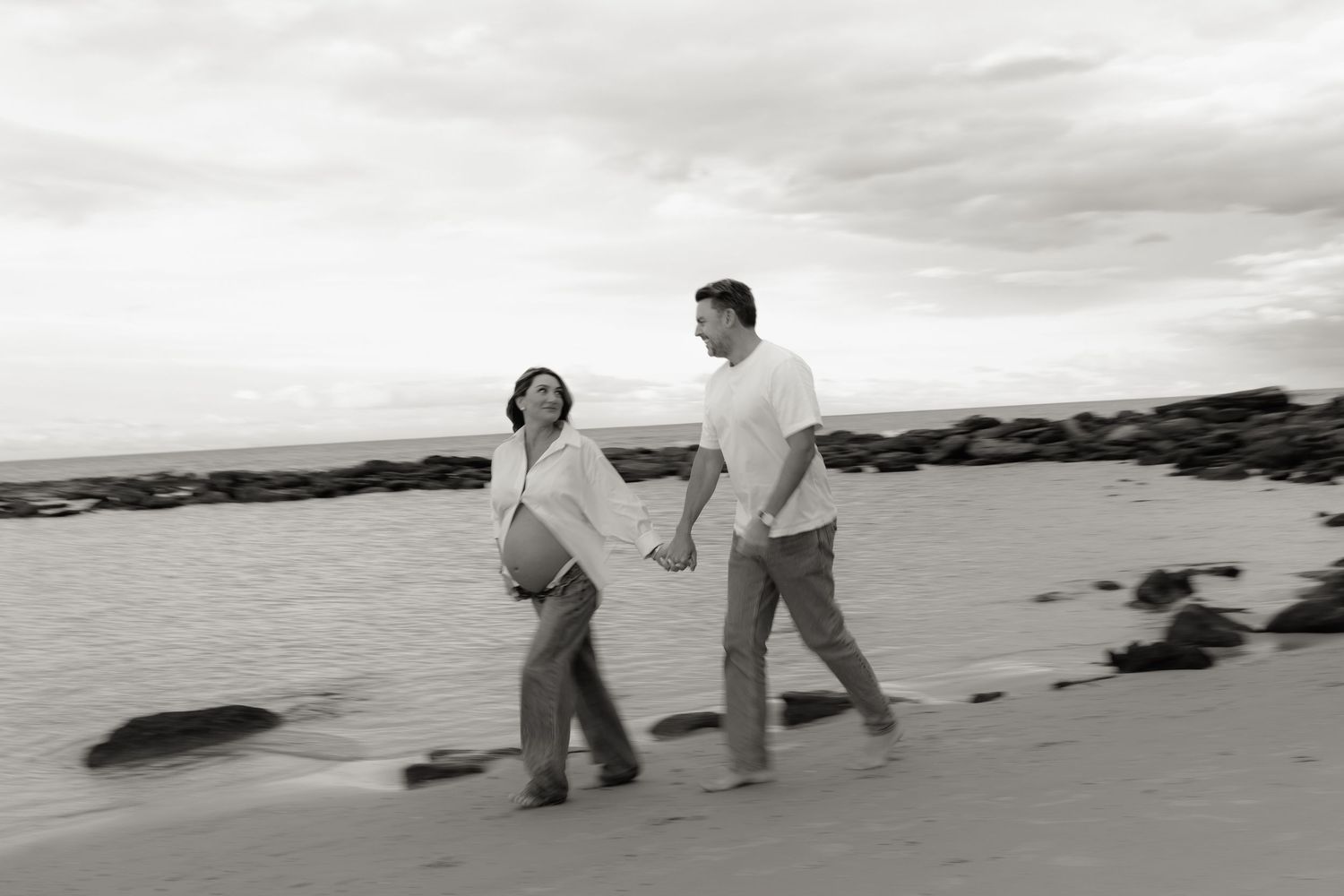 Pregnant couple holding hands walking on a beach in black and white photo.