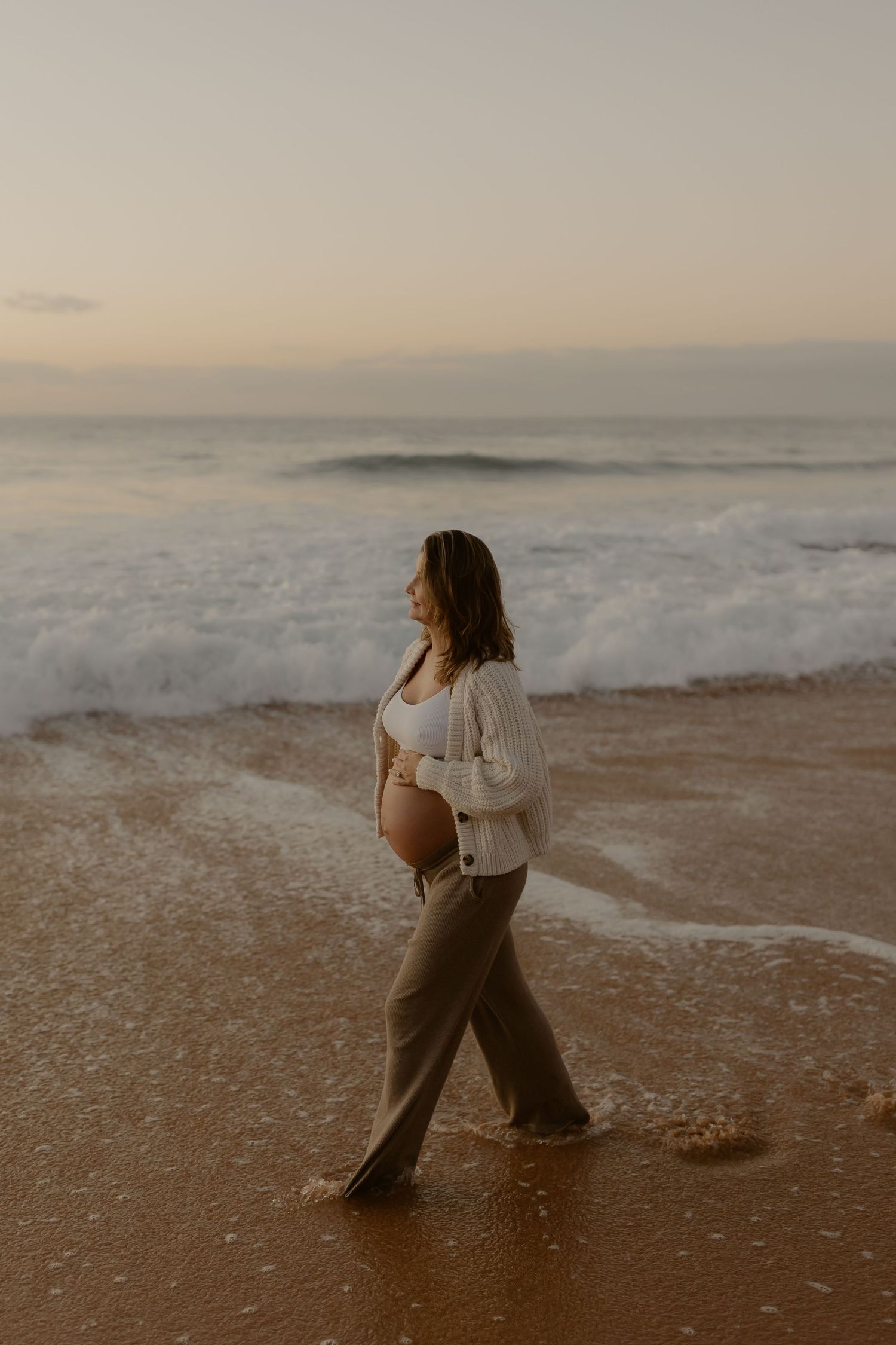 Pregnant woman walking along shoreline at sunset wearing white cardigan and olive wide-leg pants.