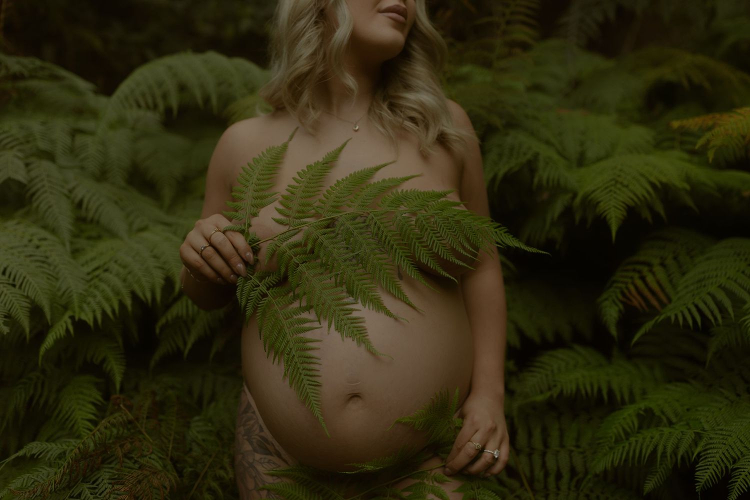 Pregnant woman posing outdoors among lush green ferns, holding a fern frond over her bare baby bump.