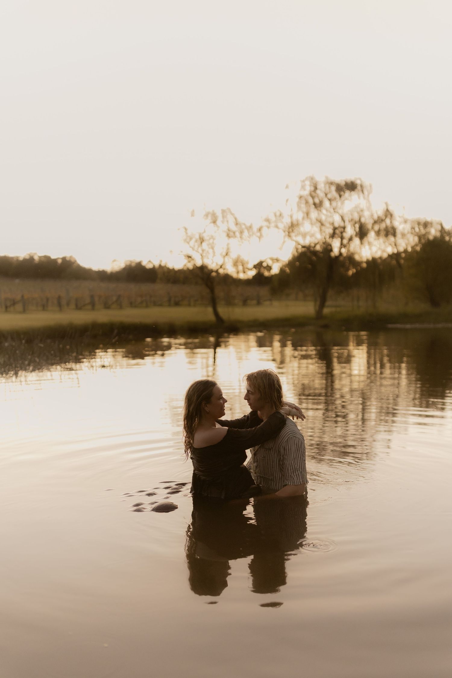 Romantic couple embracing while standing in a calm lake at golden hour with reflections on water.