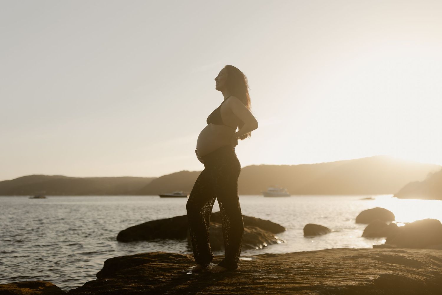 A silhouette of a pregnant woman stands on rocks by a lake at sunset, creating a peaceful maternity silhouette.