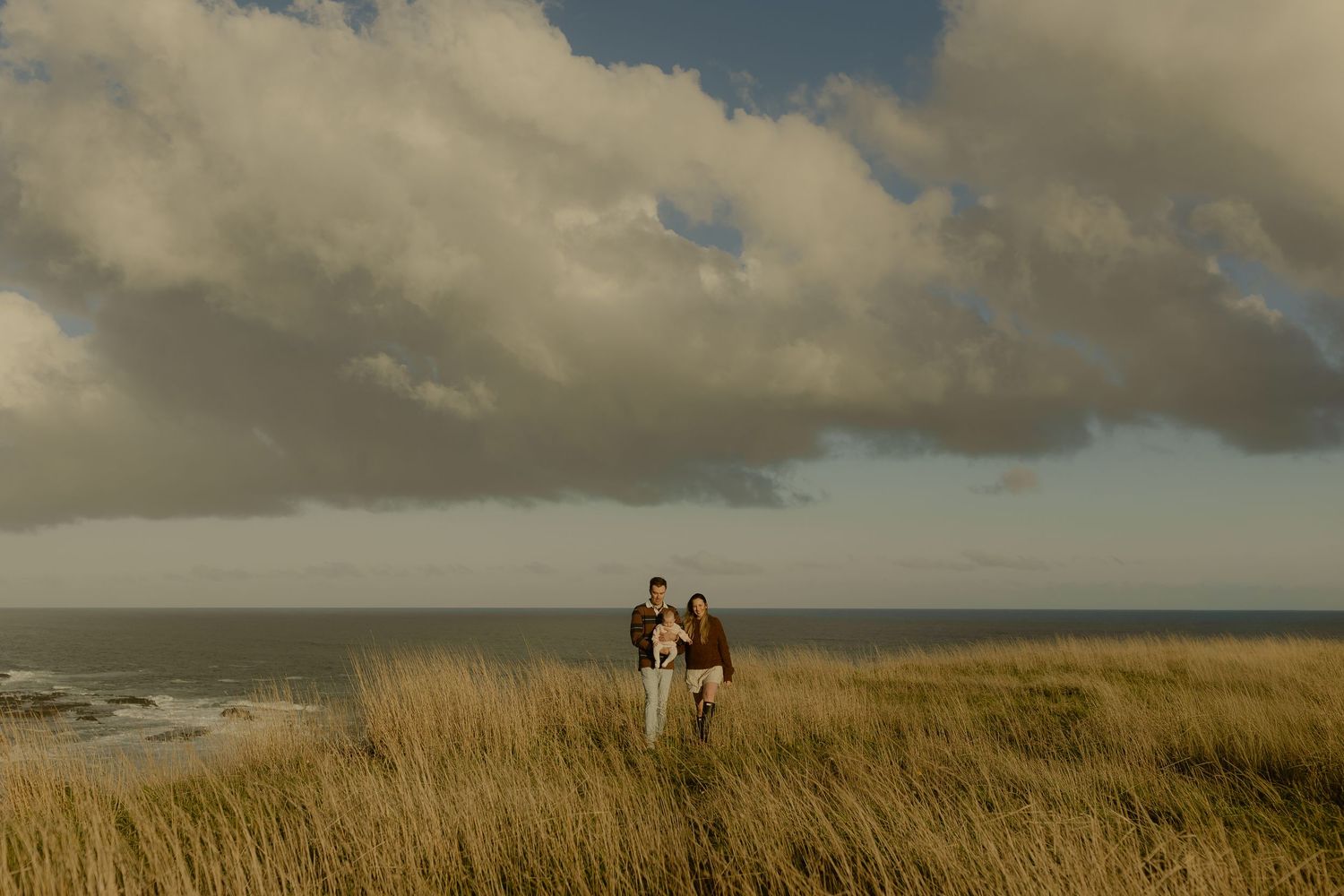 Family standing together in tall coastal grass with dramatic cloudy sky and ocean backdrop.