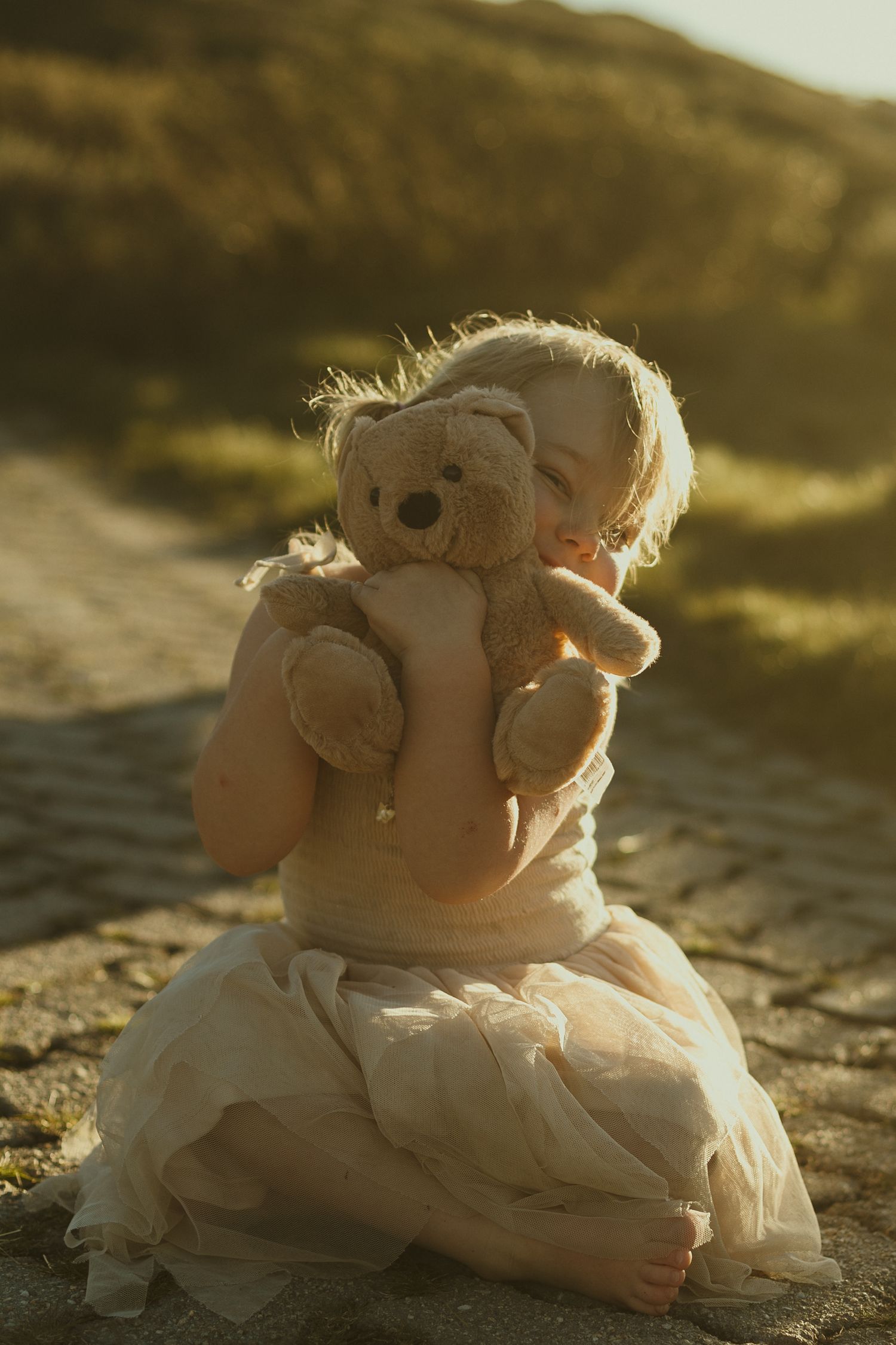 A backlit figure in a white dress sits on sand holding a teddy bear during golden hour.