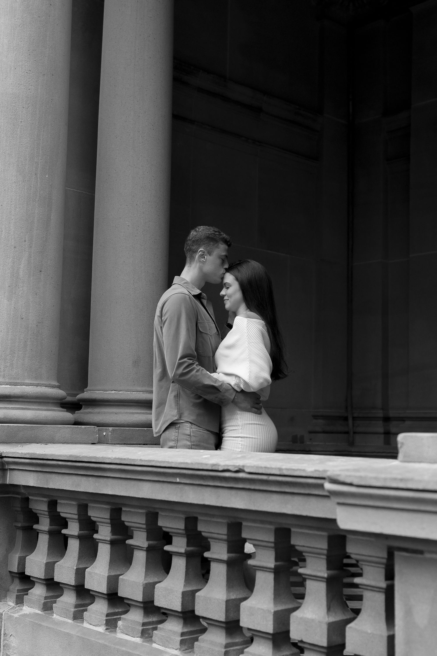 Black and white romantic portrait of a couple embracing on an architectural balcony with classic stone columns and railing.