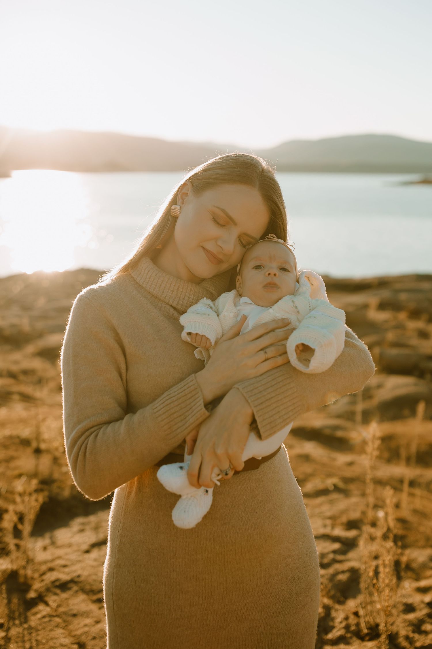 A parent wearing a tan sweater dress holds an infant outdoors during golden hour by the water's edge.