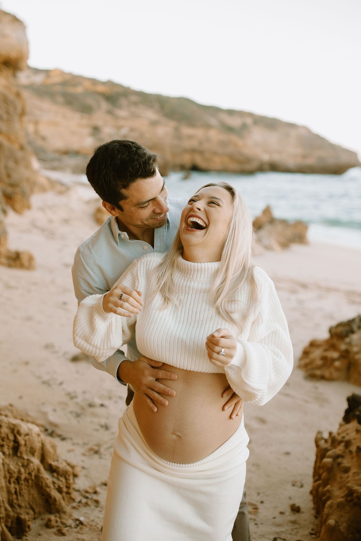 A couple shares a joyful moment together on a beach with rocky cliffs during a maternity photoshoot.