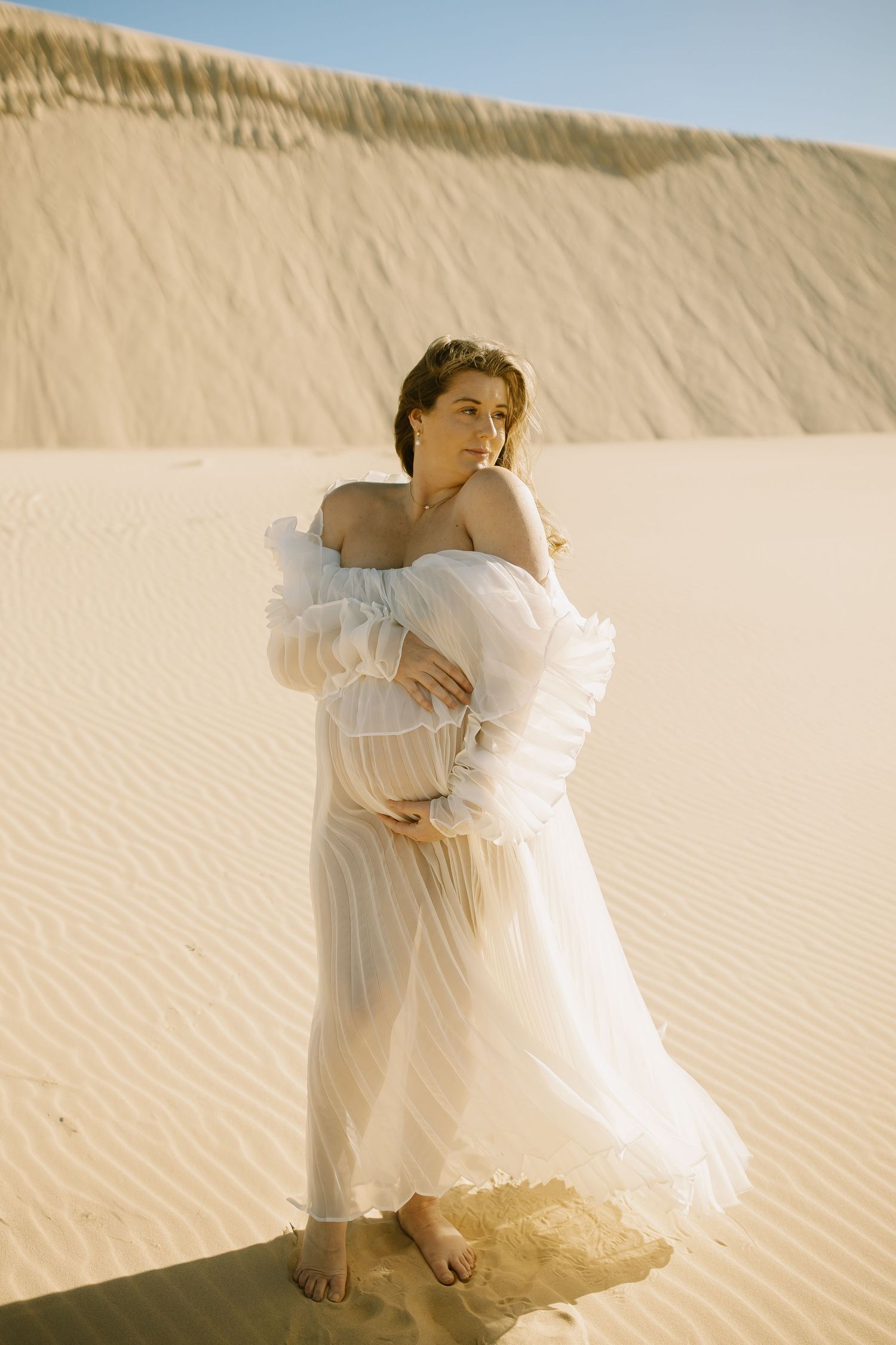A flowing white dress catches the desert breeze against sandy dunes in a dreamy ethereal portrait.