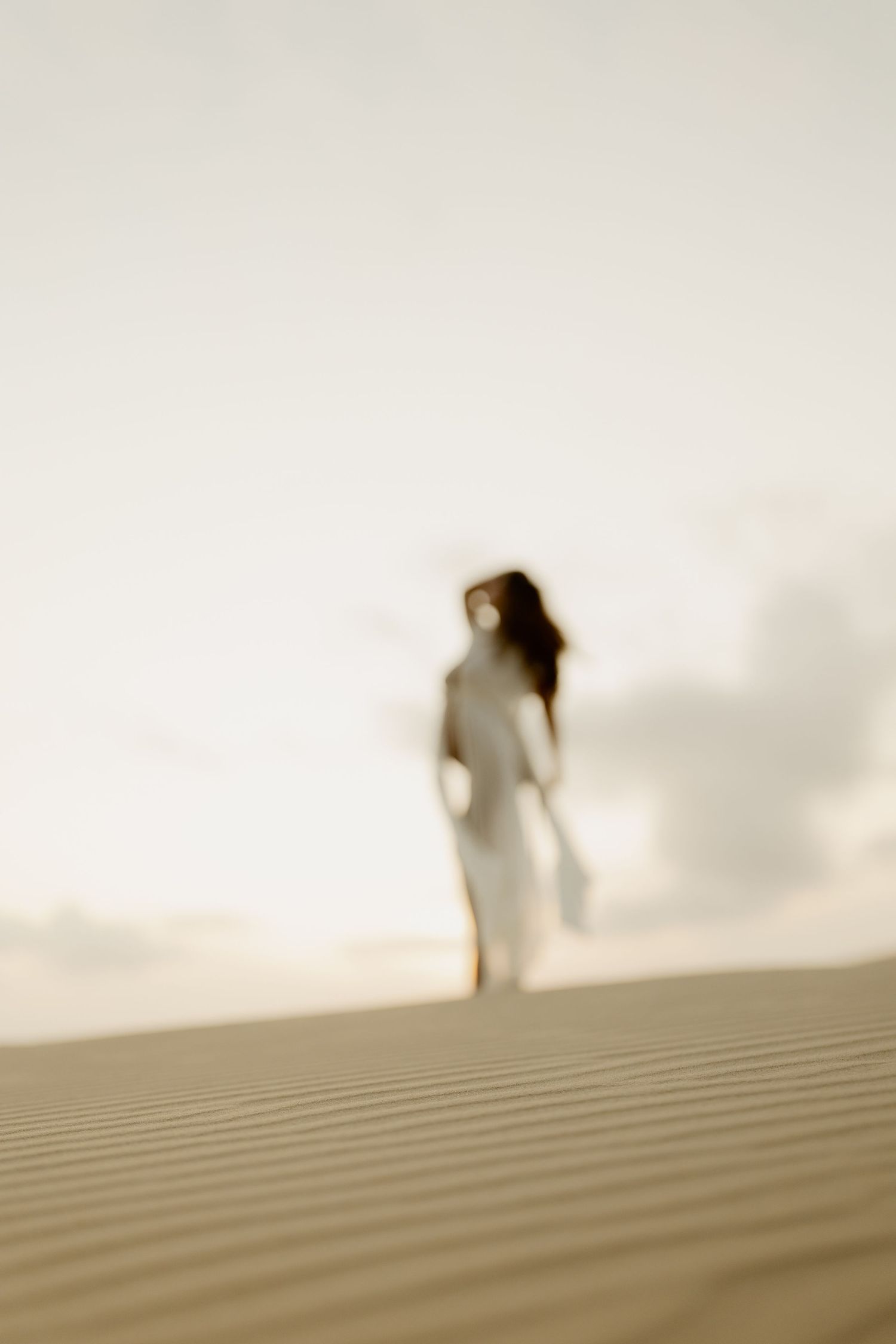 A silhouetted figure walks along the crest of a sand dune against a bright, hazy sky.