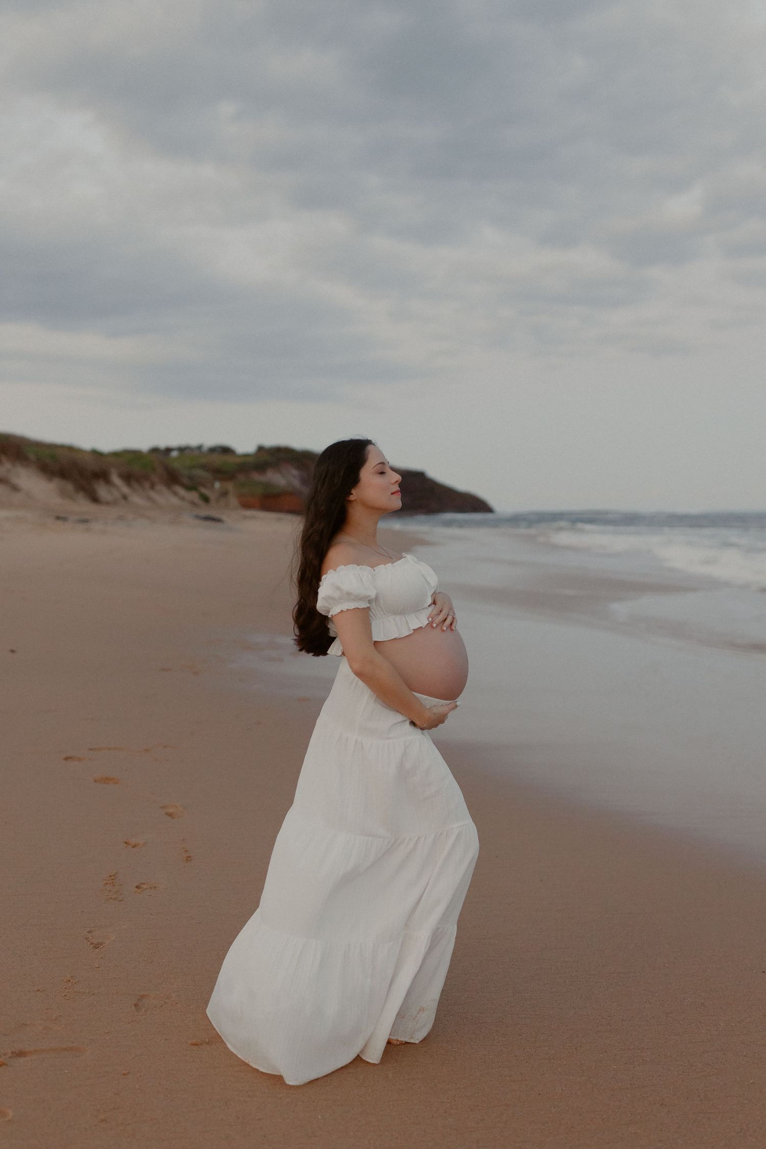 A pregnant woman in a white dress walks along a misty beach shoreline during overcast weather.