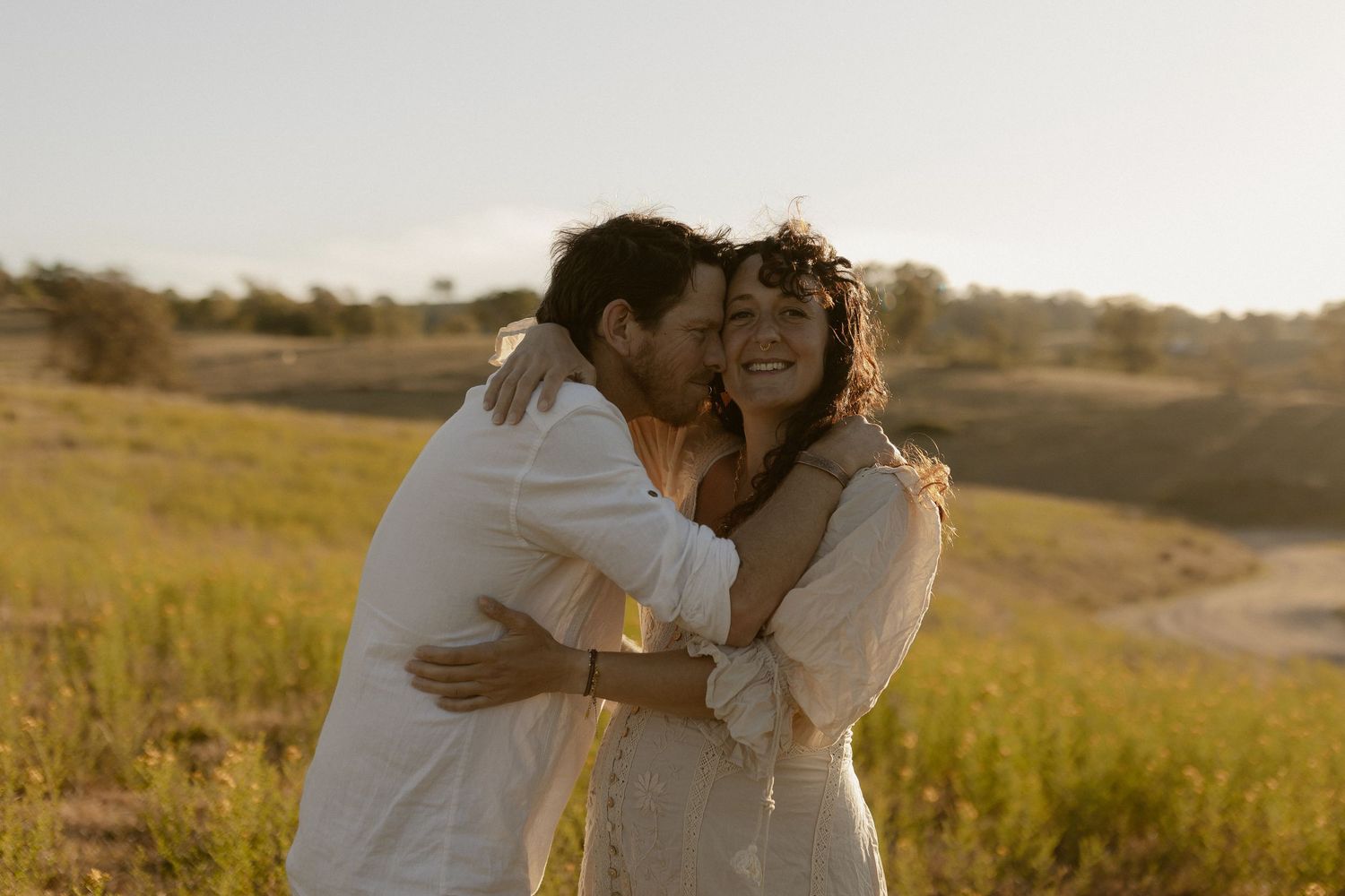 Romantic moment between couple in white outfits standing in a sunlit meadow.