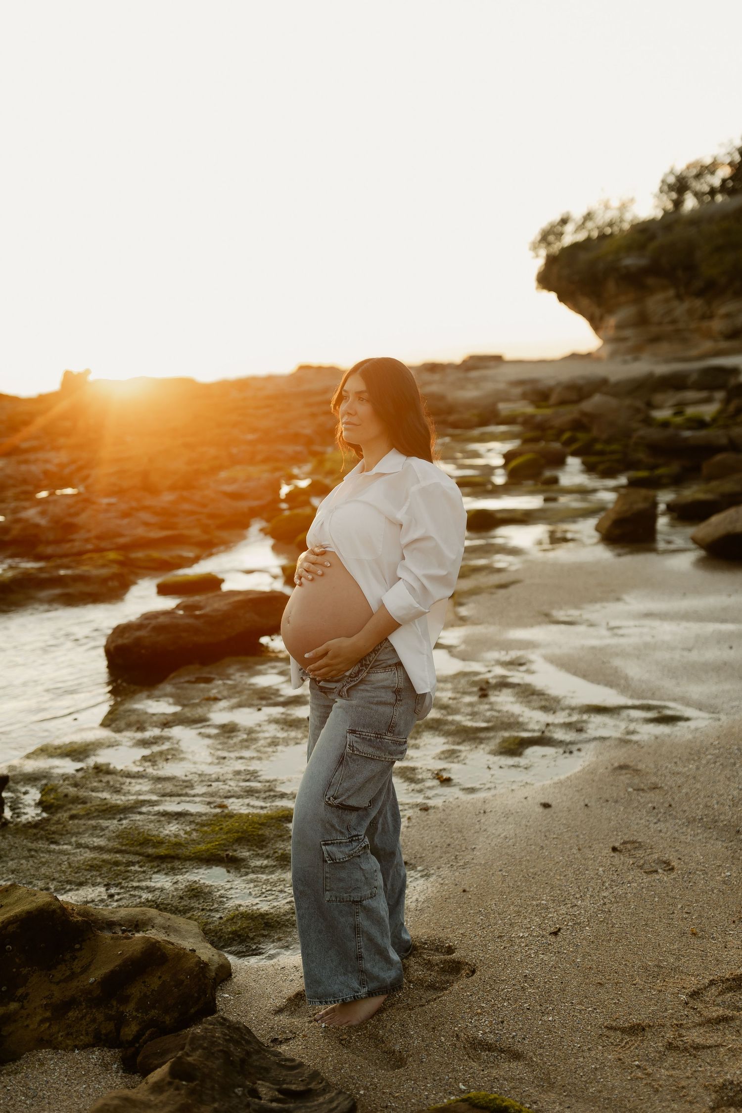 Pregnant woman in white shirt and jeans cradling baby bump on rocky beach at golden sunset.