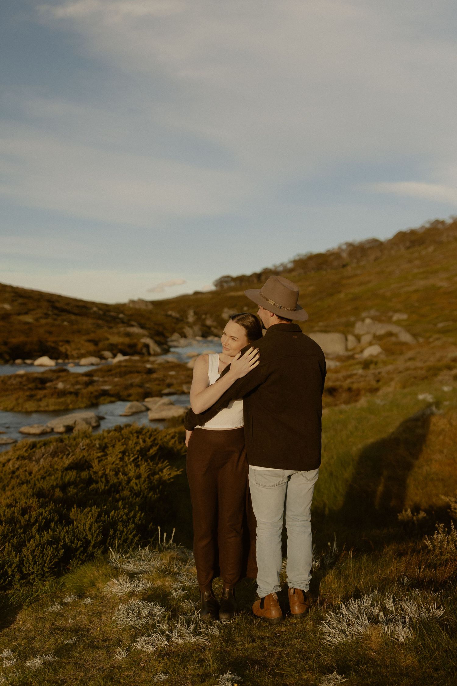 A couple shares an intimate embrace while standing on a rugged hillside with a winding stream in the background.
