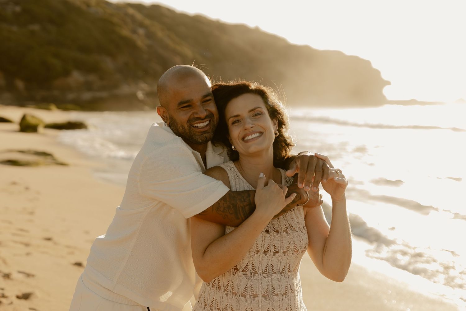 A couple shares a romantic moment on a sandy beach at sunset with ocean waves in the background.
