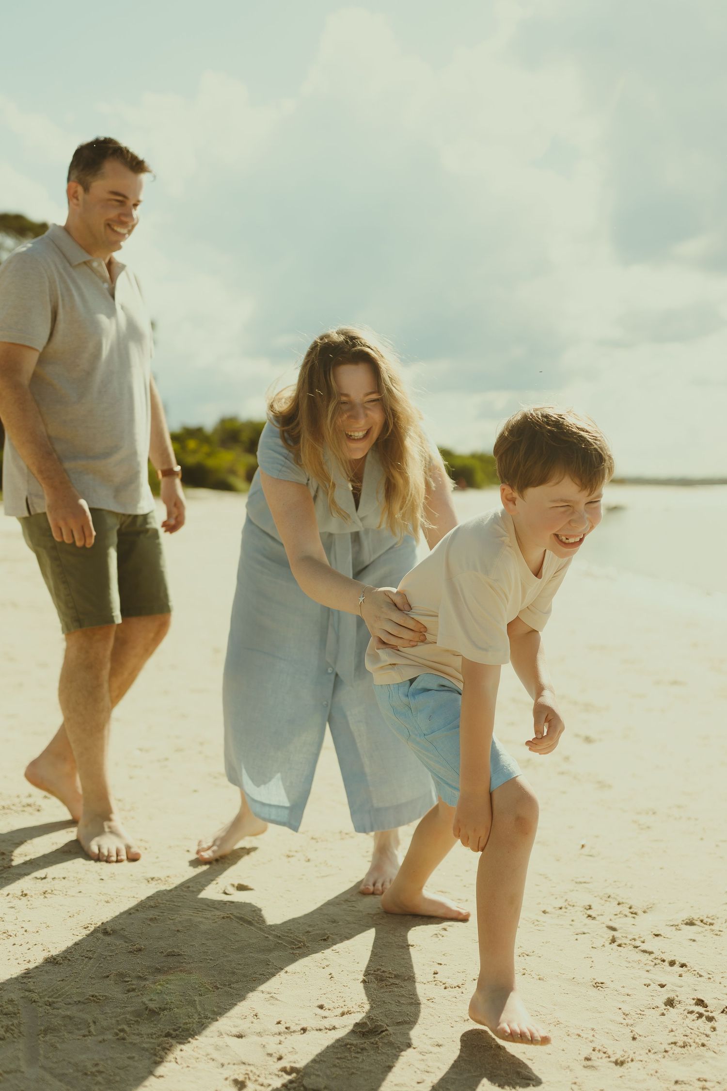 A family plays together and laughs while walking along a sunny white sand beach.