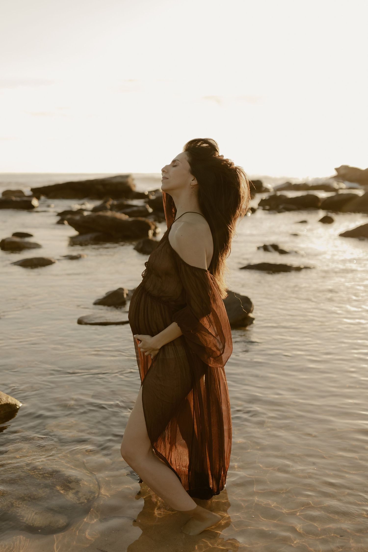 A couple embraces on a rocky beach at sunset with ocean waves in the background.