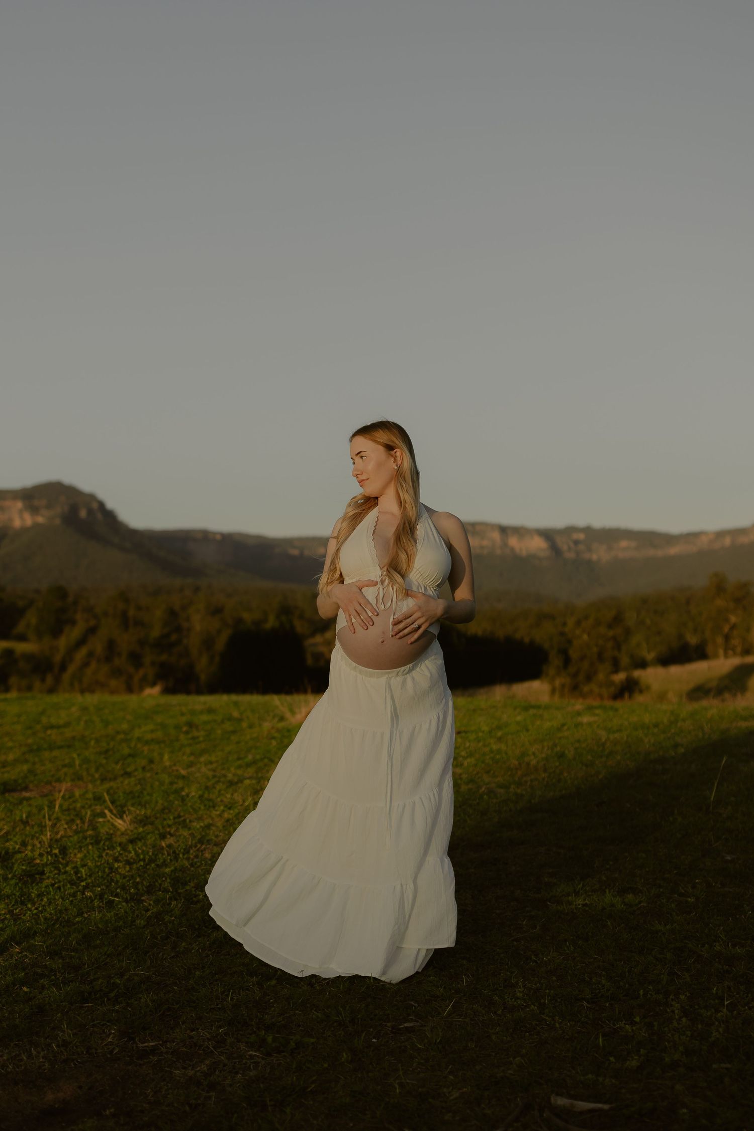 Pregnant woman in white dress holding baby bump at golden hour with mountain landscape backdrop.