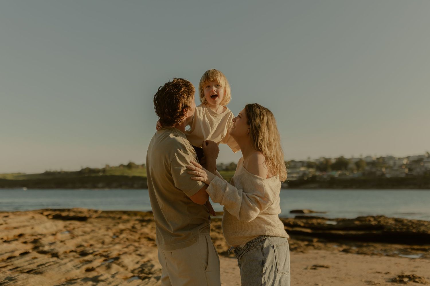 Happy family with a toddler laughing on a sandy beach in warm golden sunlight.