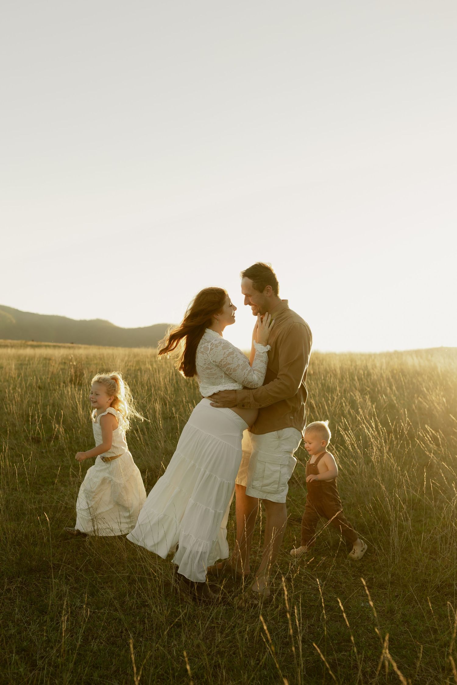 A couple embraces in a golden field at sunset while two young children play around them.