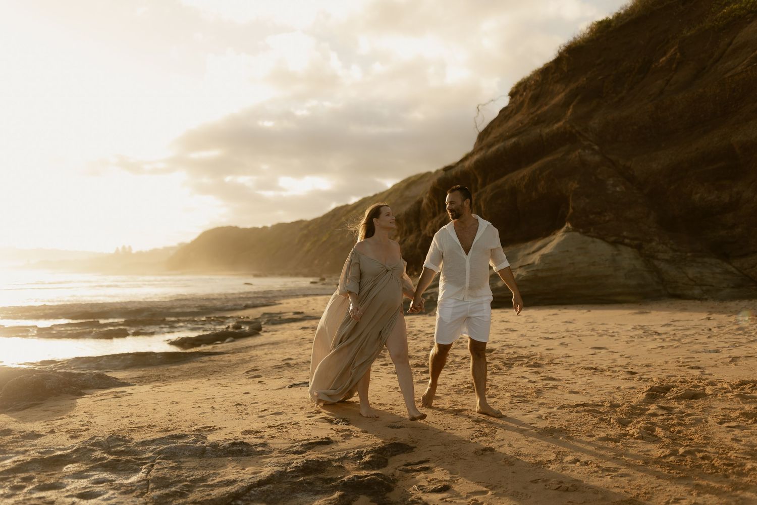 A romantic couple walks along a scenic beach at sunset with mountains and waves in the background.