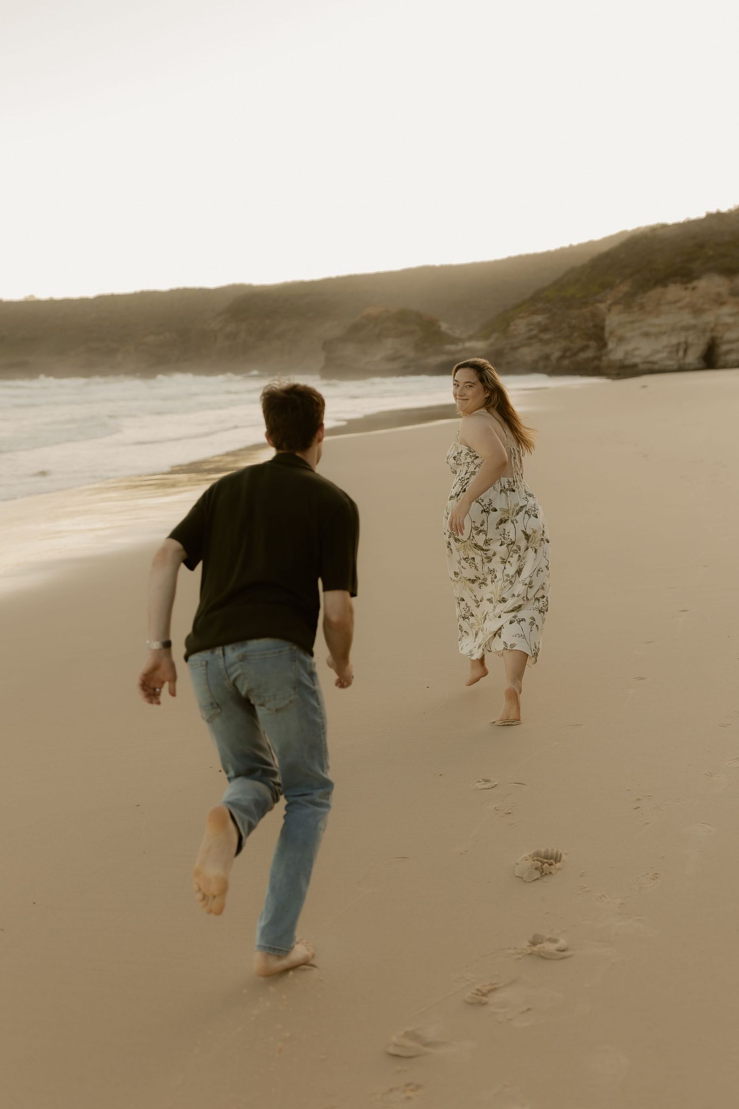 Two people walk together along a serene beach shoreline leaving footprints in the sand at sunset.