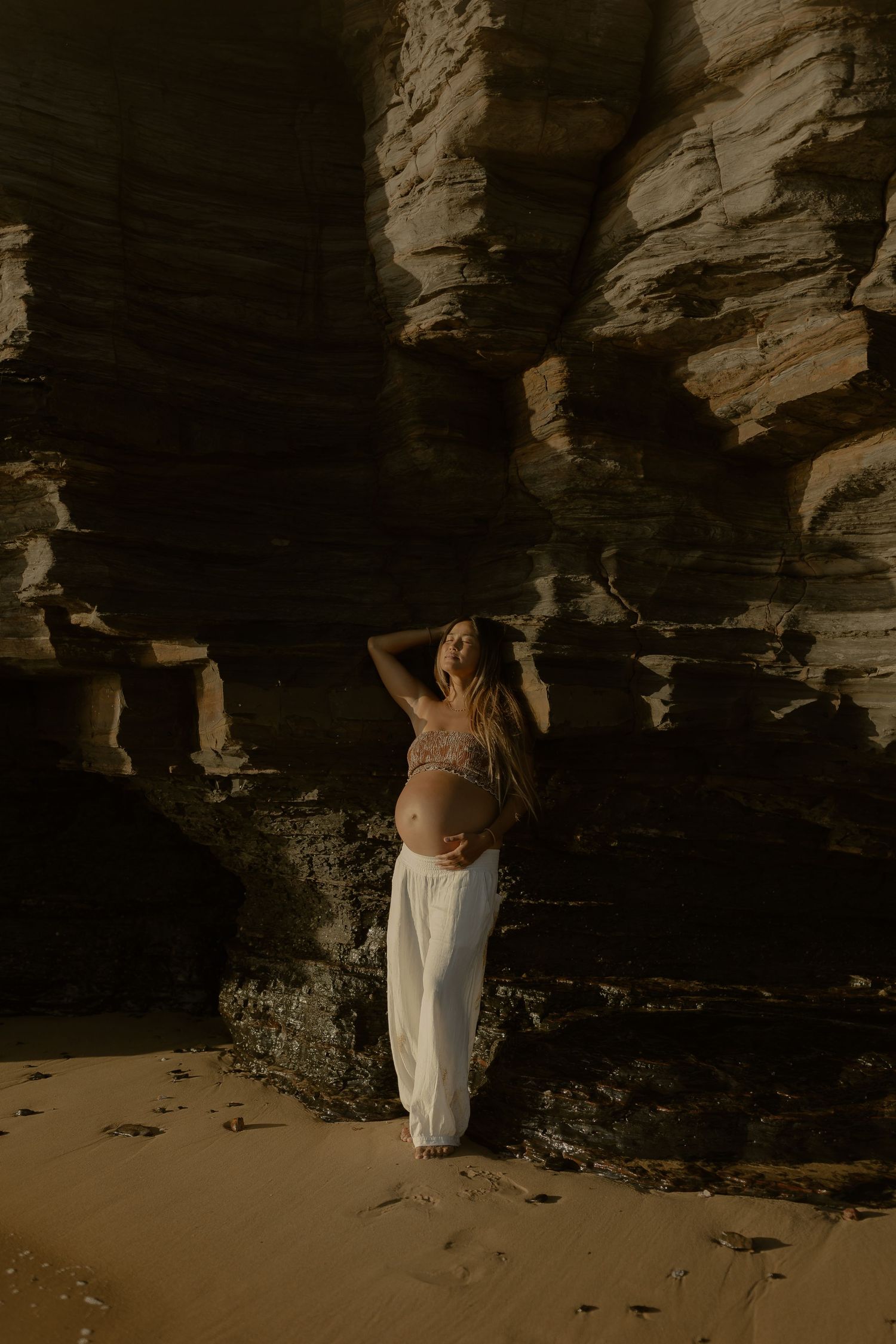 Figure in flowing white dress poses dramatically against rocky coastal cave formations at sunset.