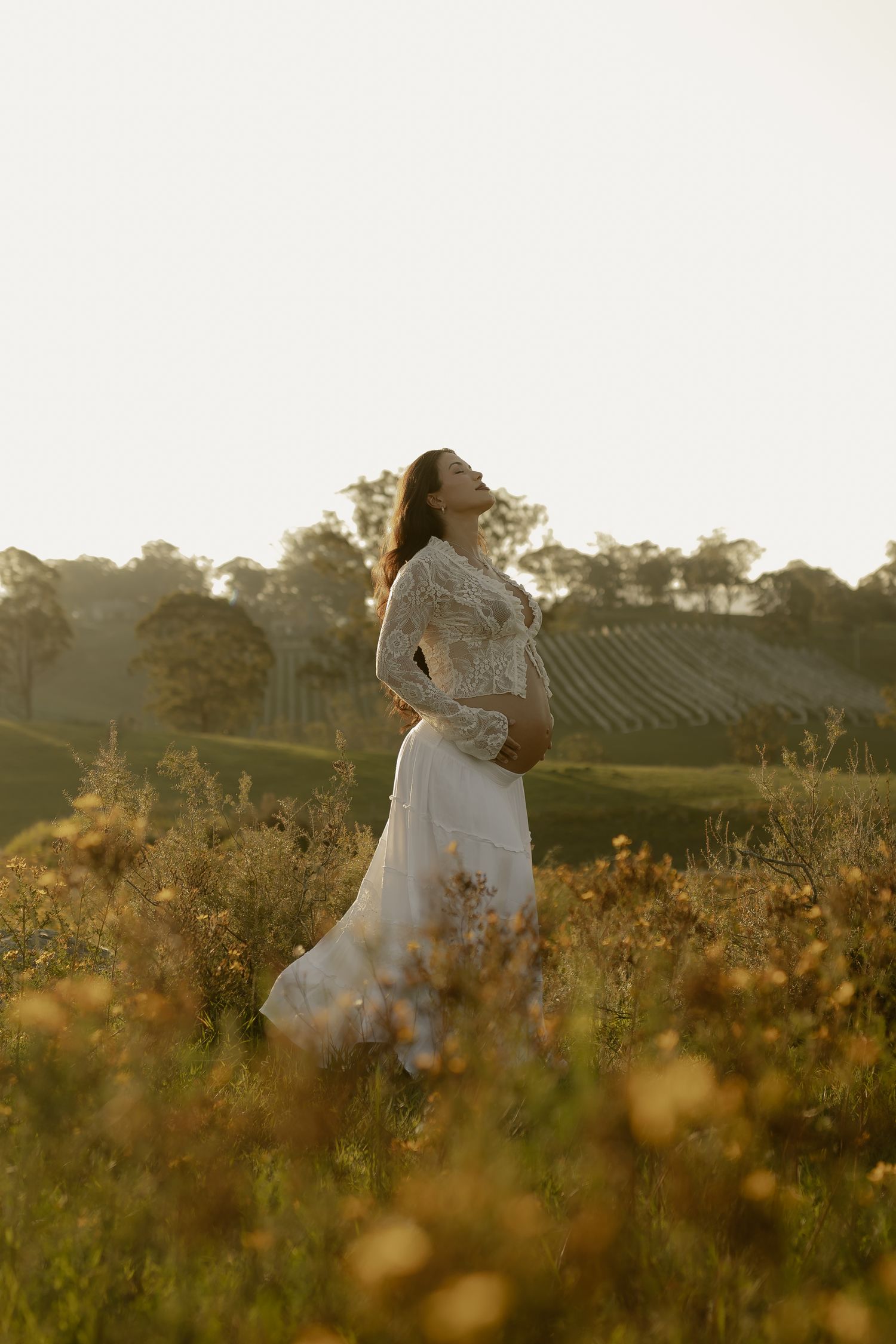 Pregnant woman in lace dress poses elegantly in golden wildflower field at sunset.