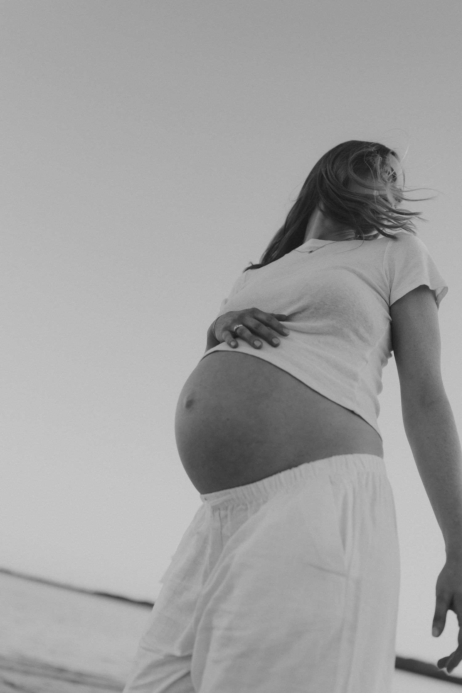 Black and white photo of pregnant woman in white outfit holding her bare baby bump against clear sky.