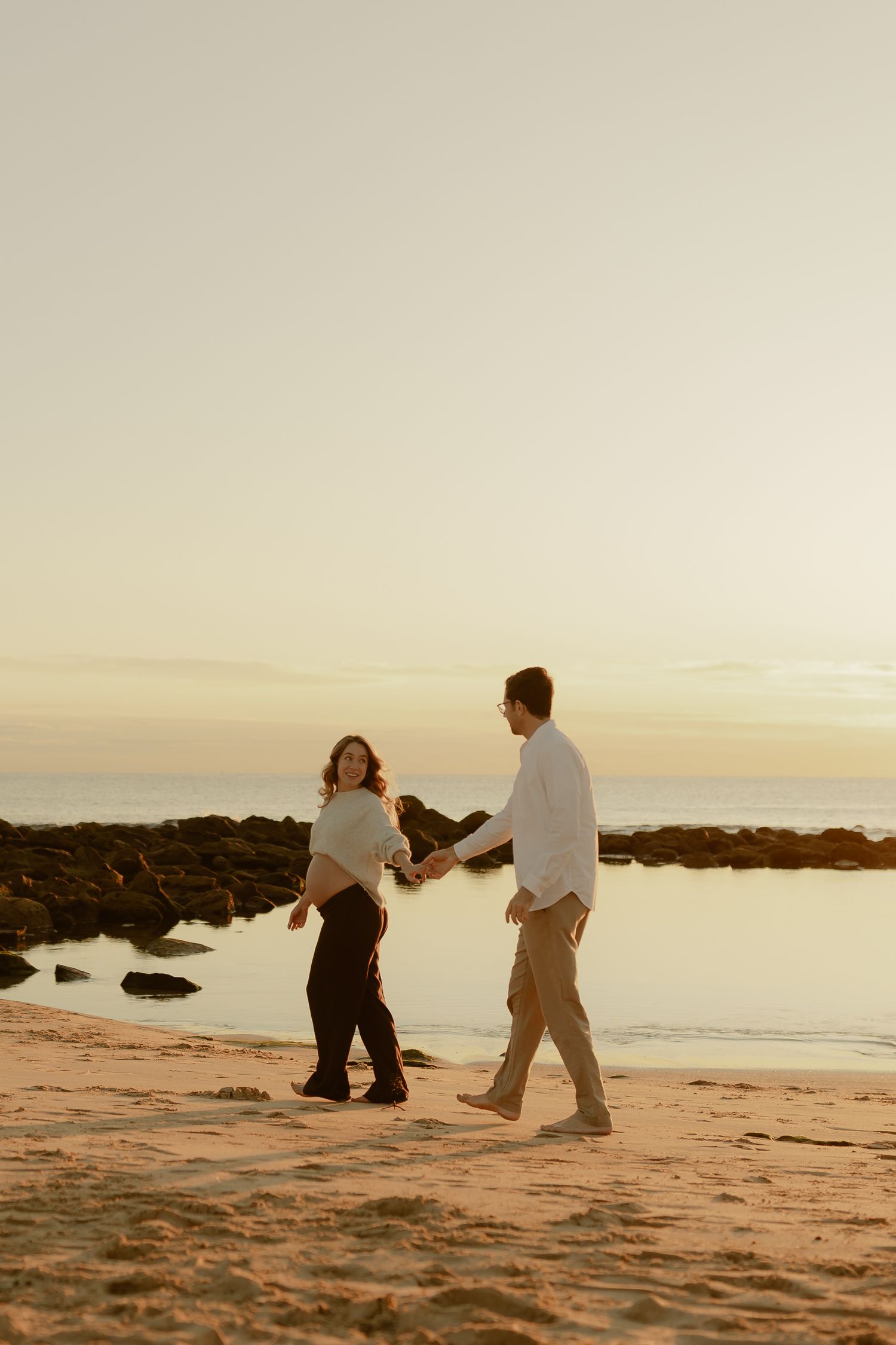 Couple holding hands walking along sandy beach at sunset with rocky coastline in background.