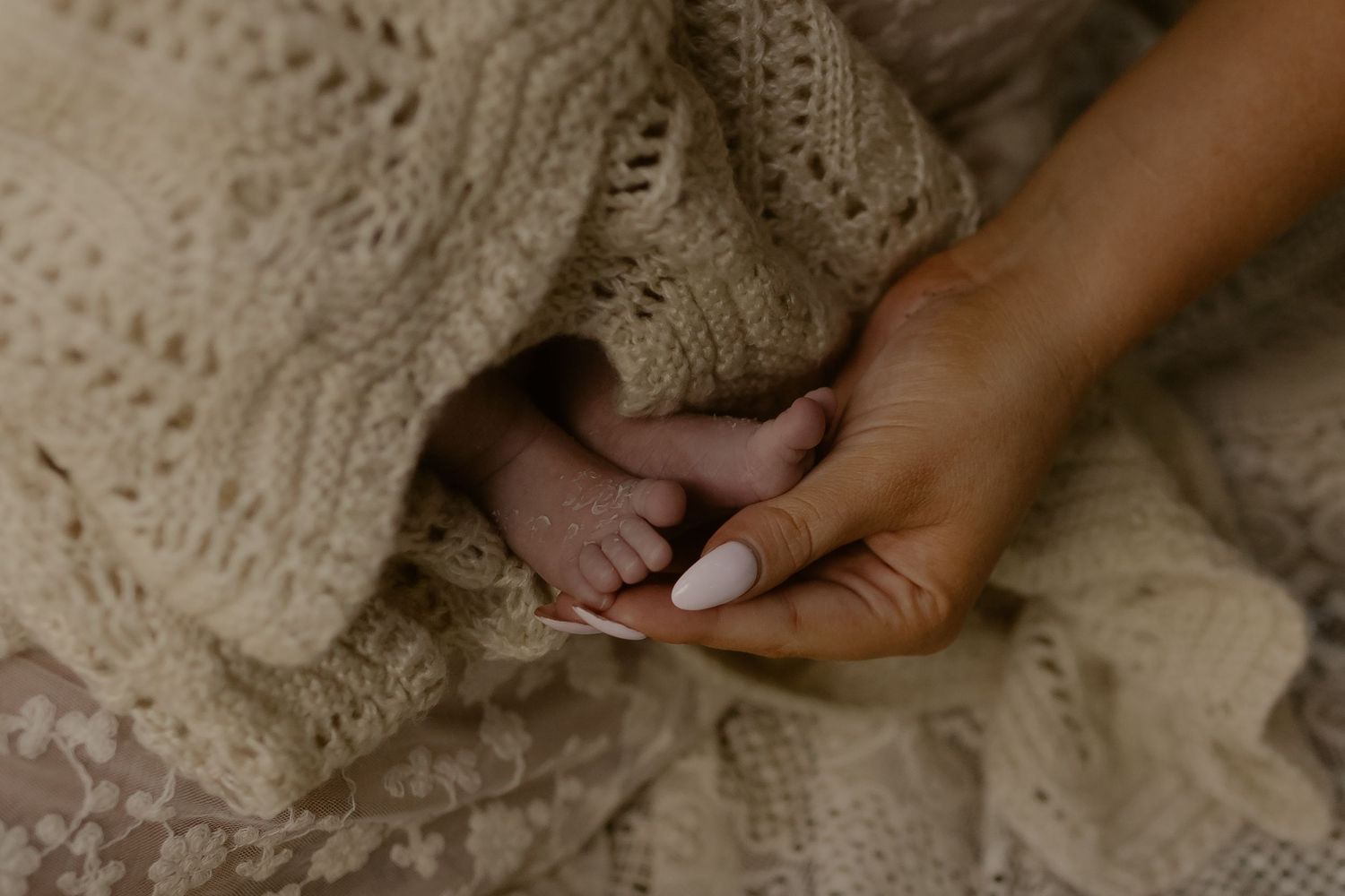 A mother's hand with white manicured nails gently holds tiny newborn baby feet wrapped in a soft cream knit blanket.
