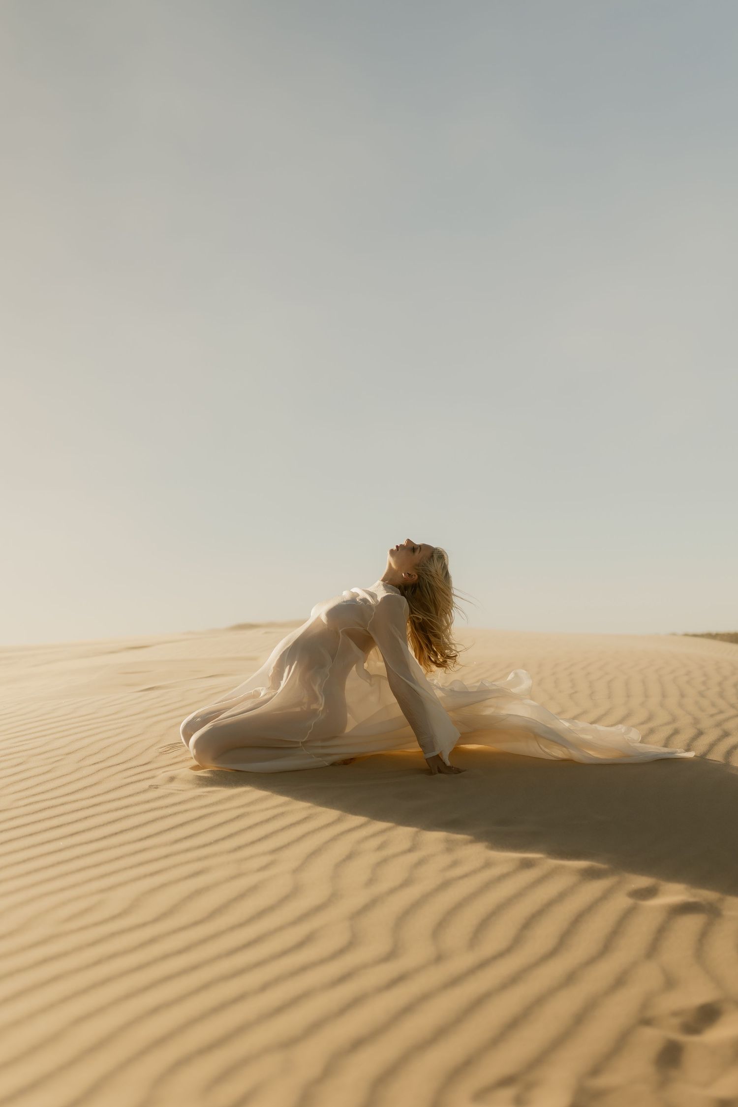 Woman in flowing white dress kneeling gracefully on golden sand dunes in a vast desert landscape.