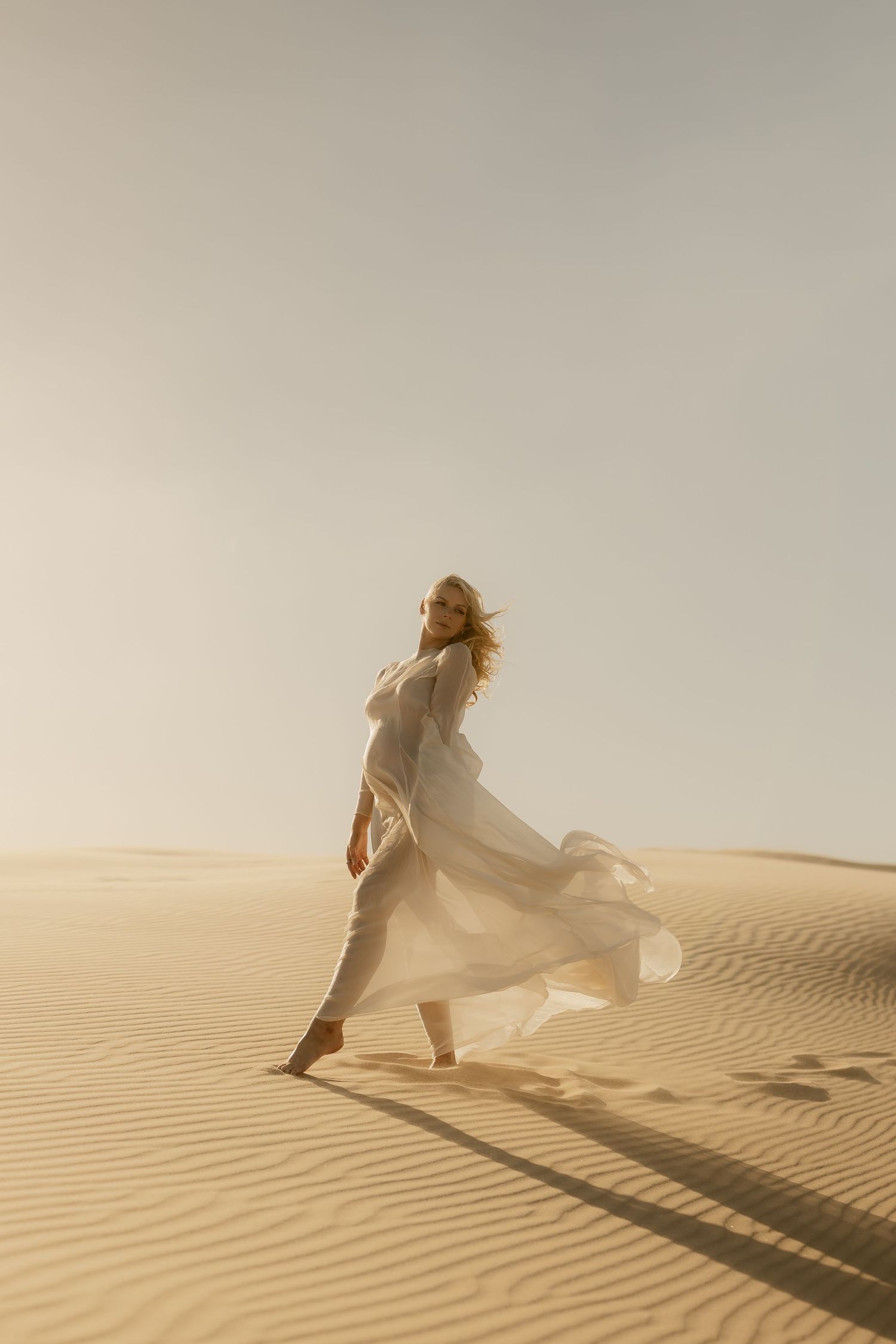 Pregnant woman in flowing white dress walking on desert sand dunes with wind blowing fabric.