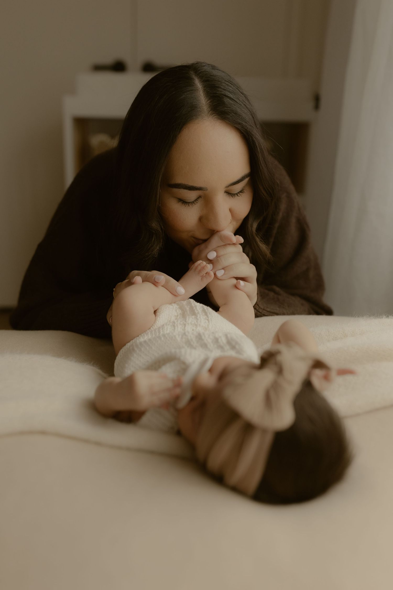 A mother tenderly kisses her newborn baby's tiny feet while lying on a soft white bed.