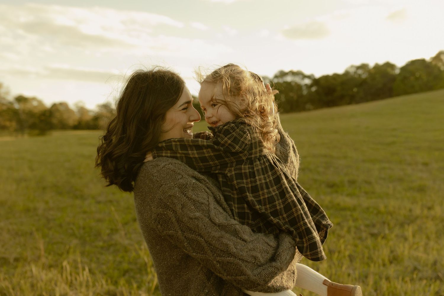Mother holding young child in warm embrace in a sunlit green field at golden hour.