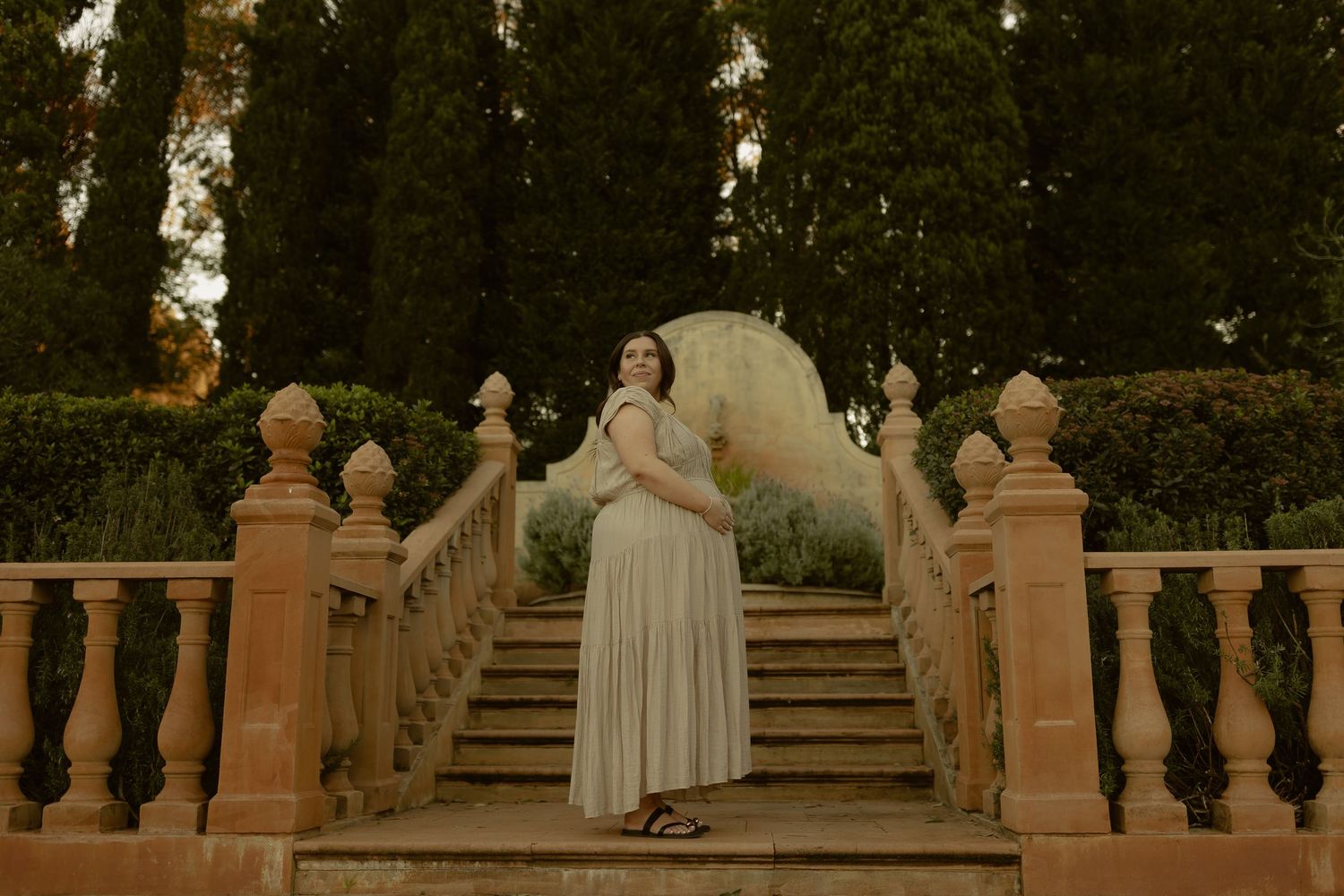 Pregnant woman in white dress poses on ornate stone staircase surrounded by cypress trees in a garden.