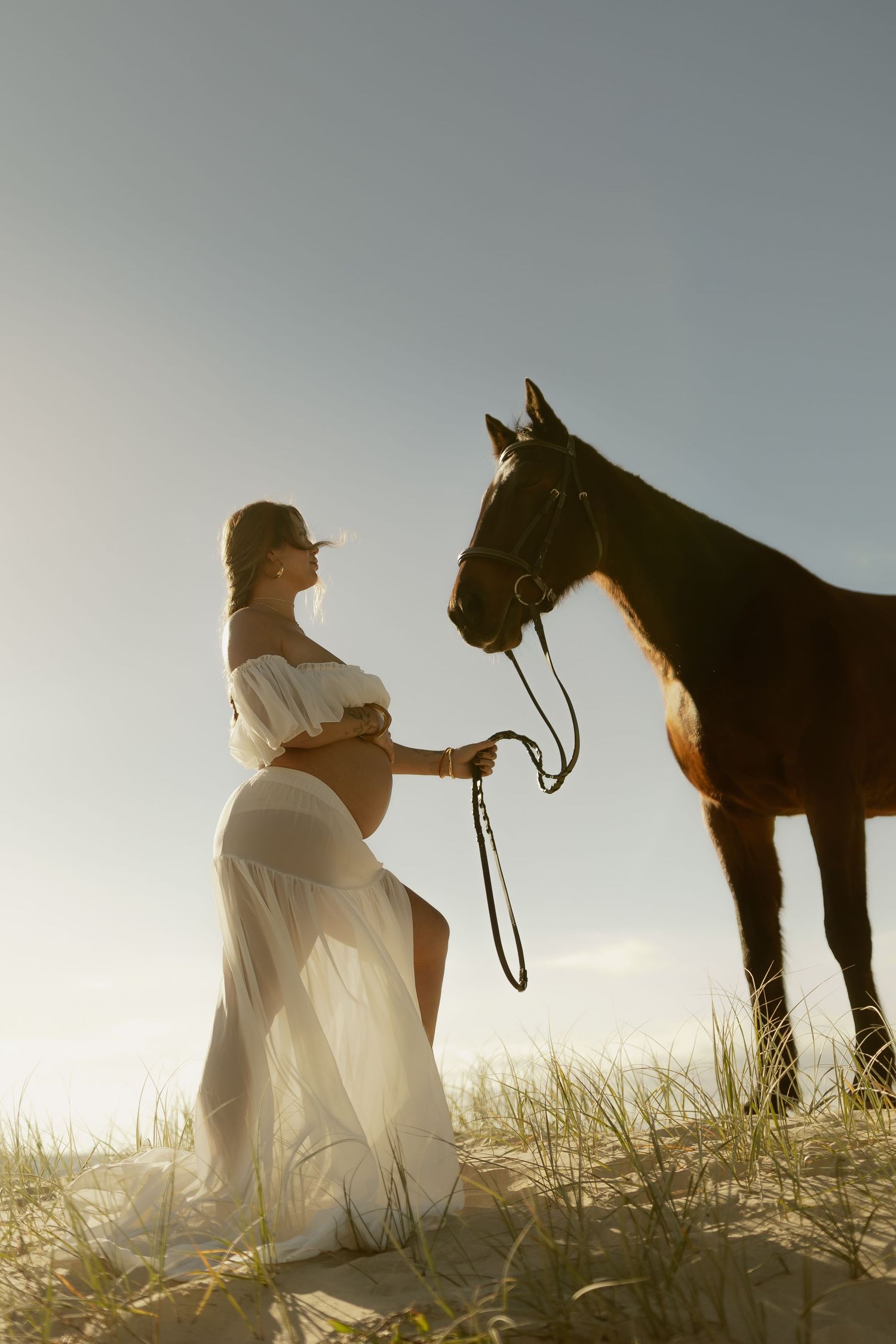 Pregnant woman in white dress kneels beside brown horse on sandy dunes at sunset.