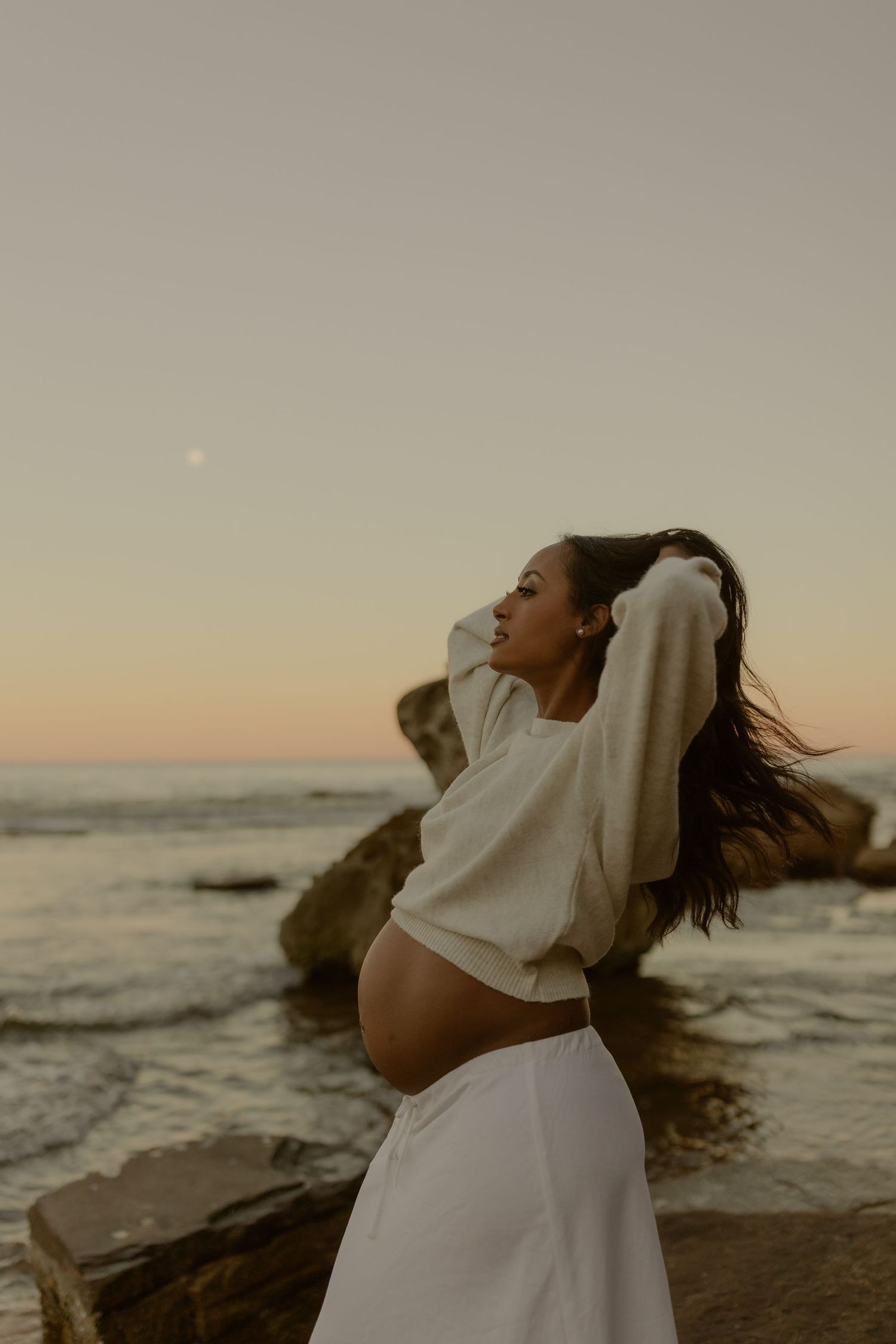 Pregnant woman in white crop top and skirt poses on rocks at sunset beach, cradling bump with wind in hair.